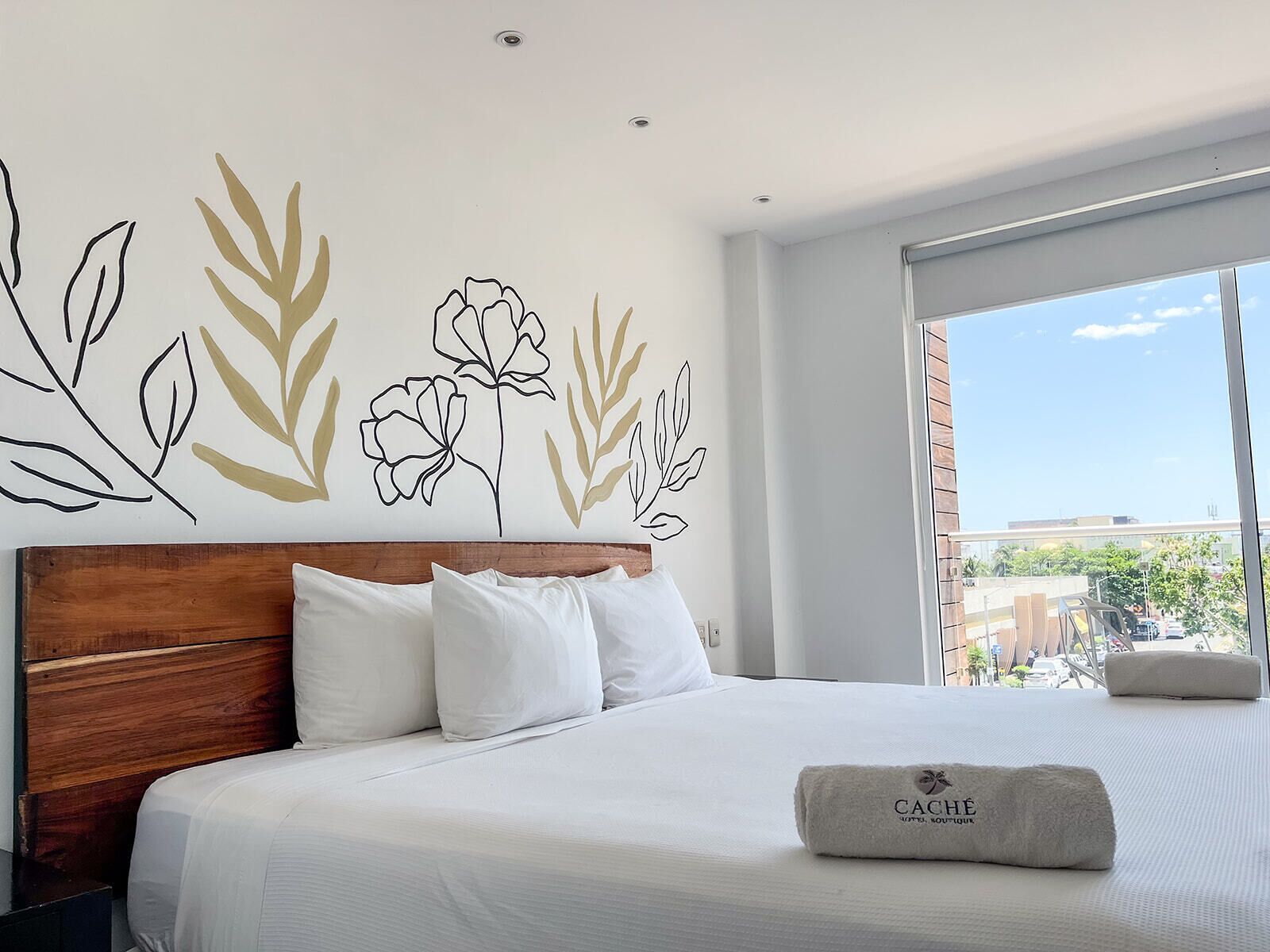 Une chambre d’hôtel avec une tête de lit en bois, du linge de lit blanc et une grande fenêtre donnant sur la ville.