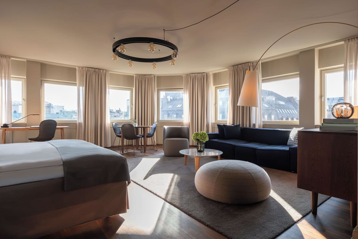 A modern hotel room with a bed, sofa, ottoman, and a dining area with chairs and a table.