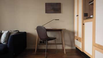 Una habitación con un sofá de color oscuro, un escritorio de madera con una silla, un pequeño marco en la pared y un armario con espejo.