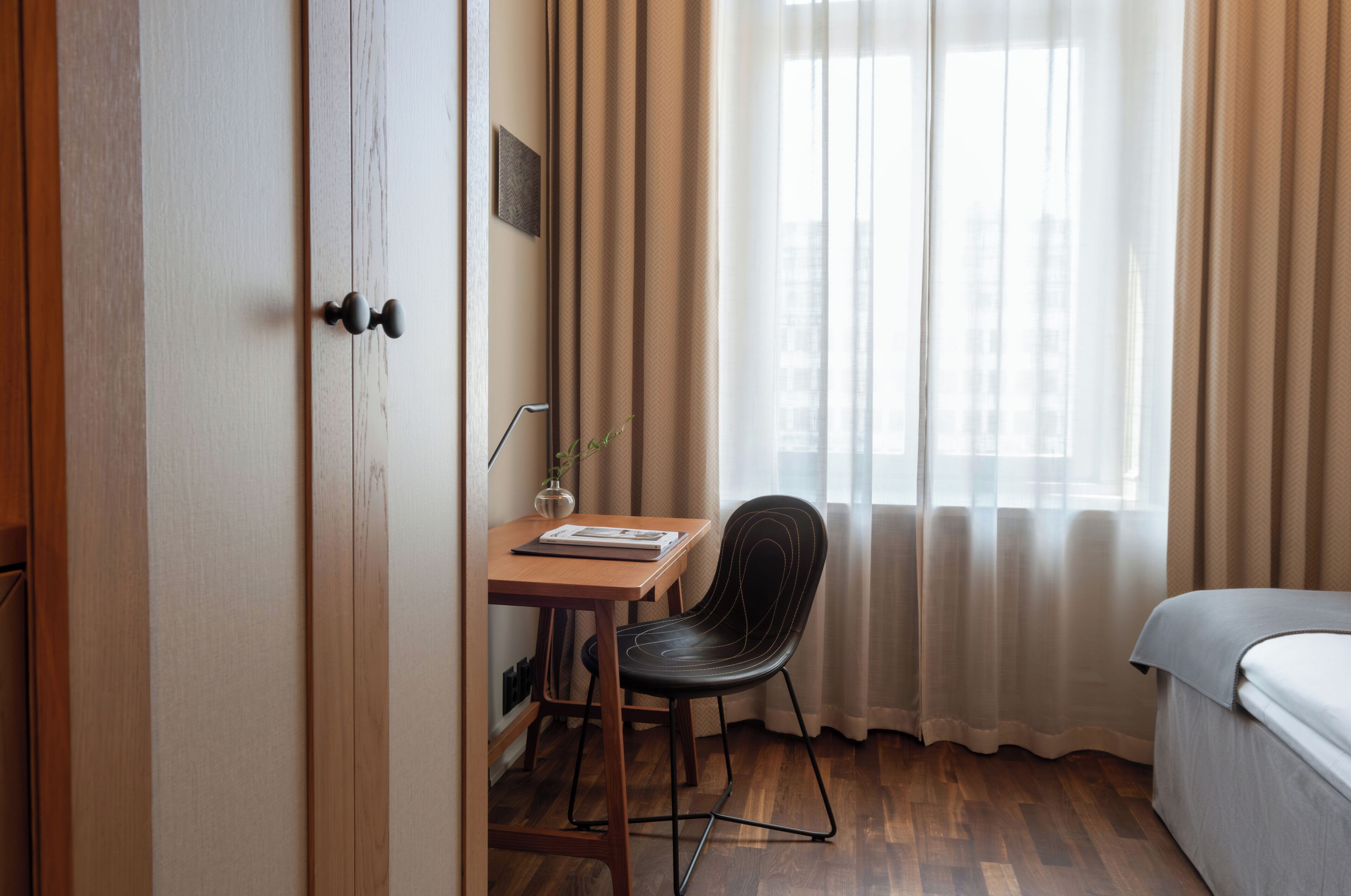 A hotel room with a wooden desk, a chair, a bed, and a window with curtains.