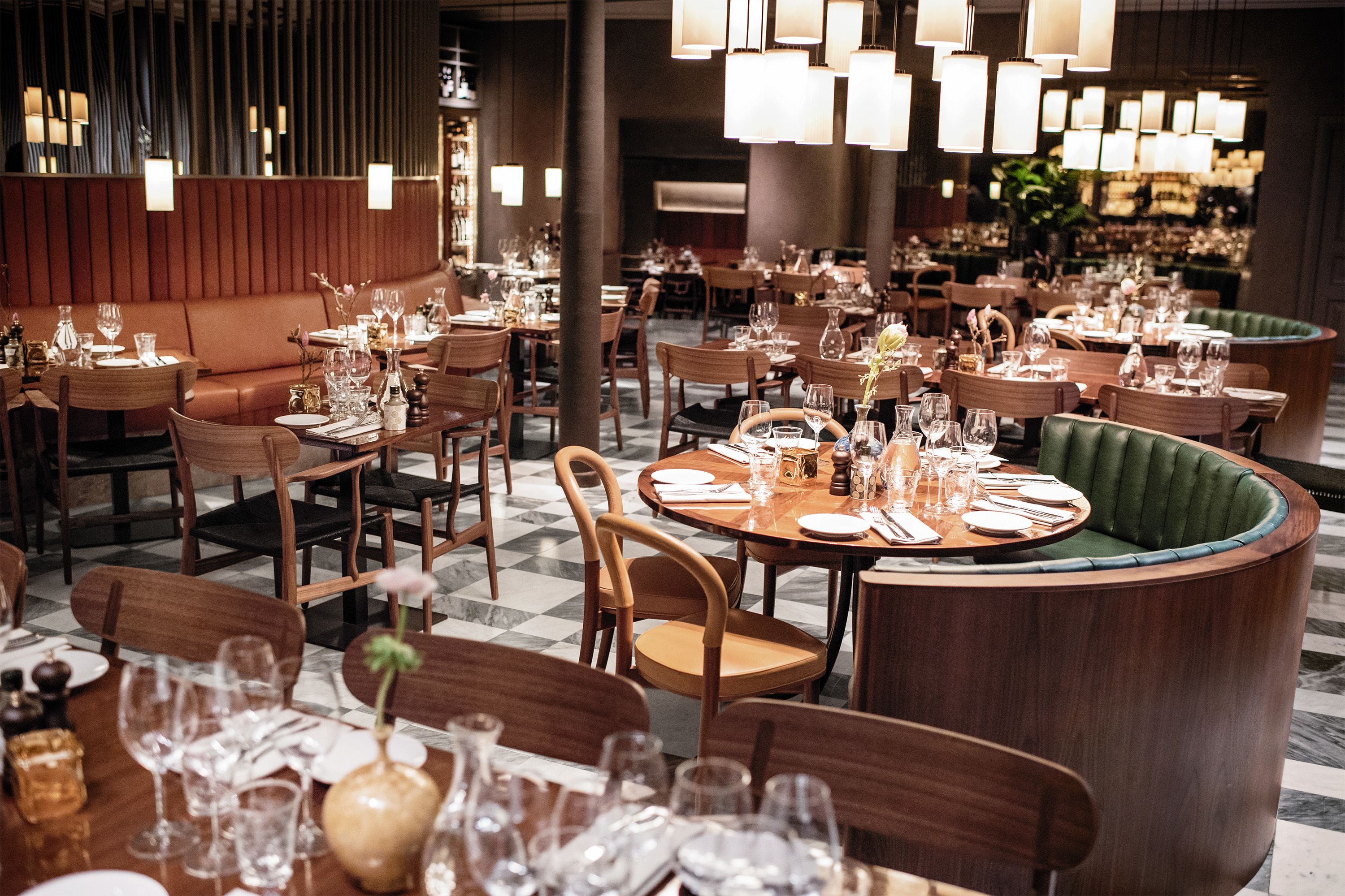 A restaurant with multiple dining tables set with fine china, glasses, and cutlery. There are wooden chairs and a green upholstered booth. The room has a modern design with pendant lighting.