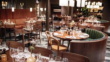 En restaurant med flere spiseborde dækket med porcelæn, glas og bestik. Træstole og en grøn polstret bænk. Rummet har et moderne design med hængende lamper.