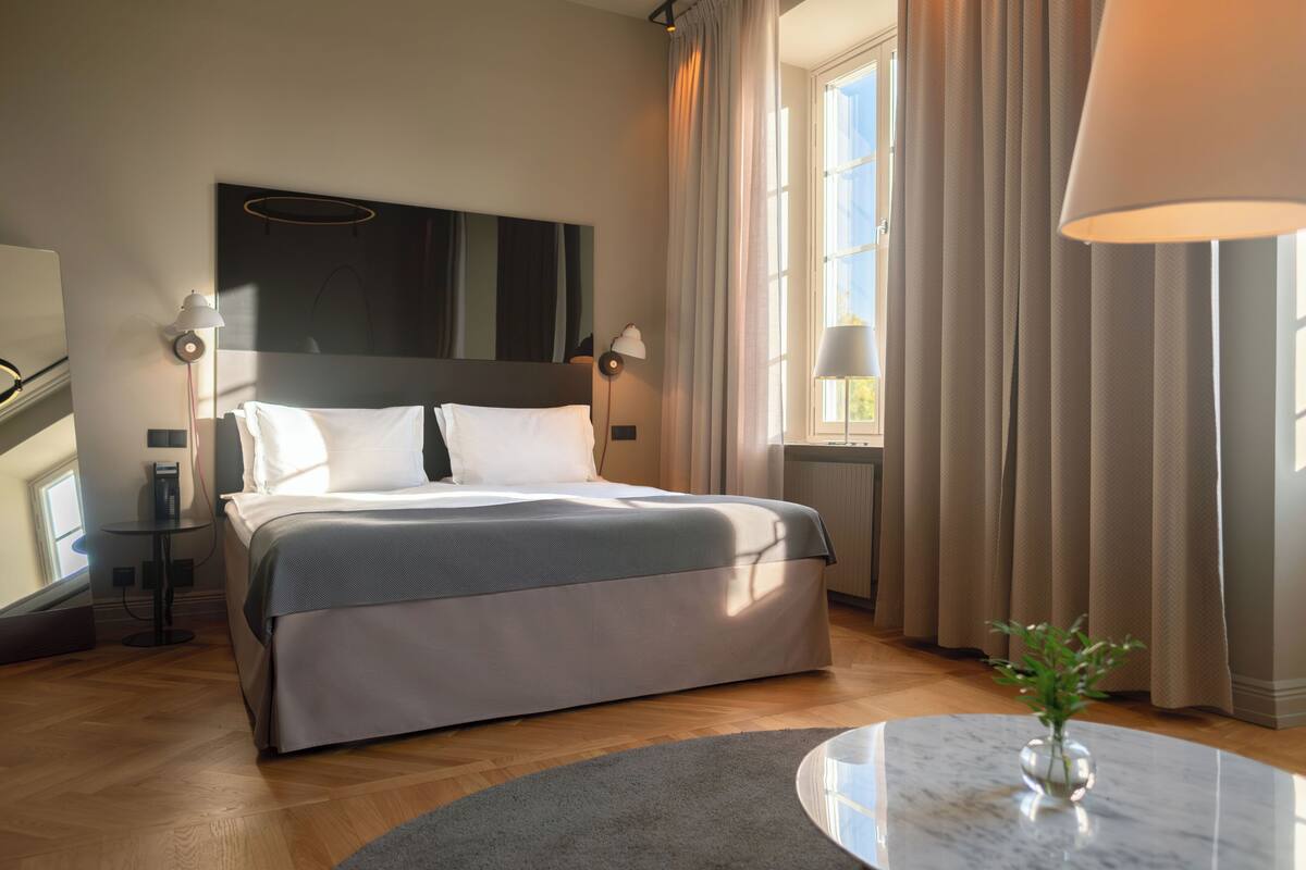 A modern hotel room with a large bed, a round marble table, and a view of the outdoors.