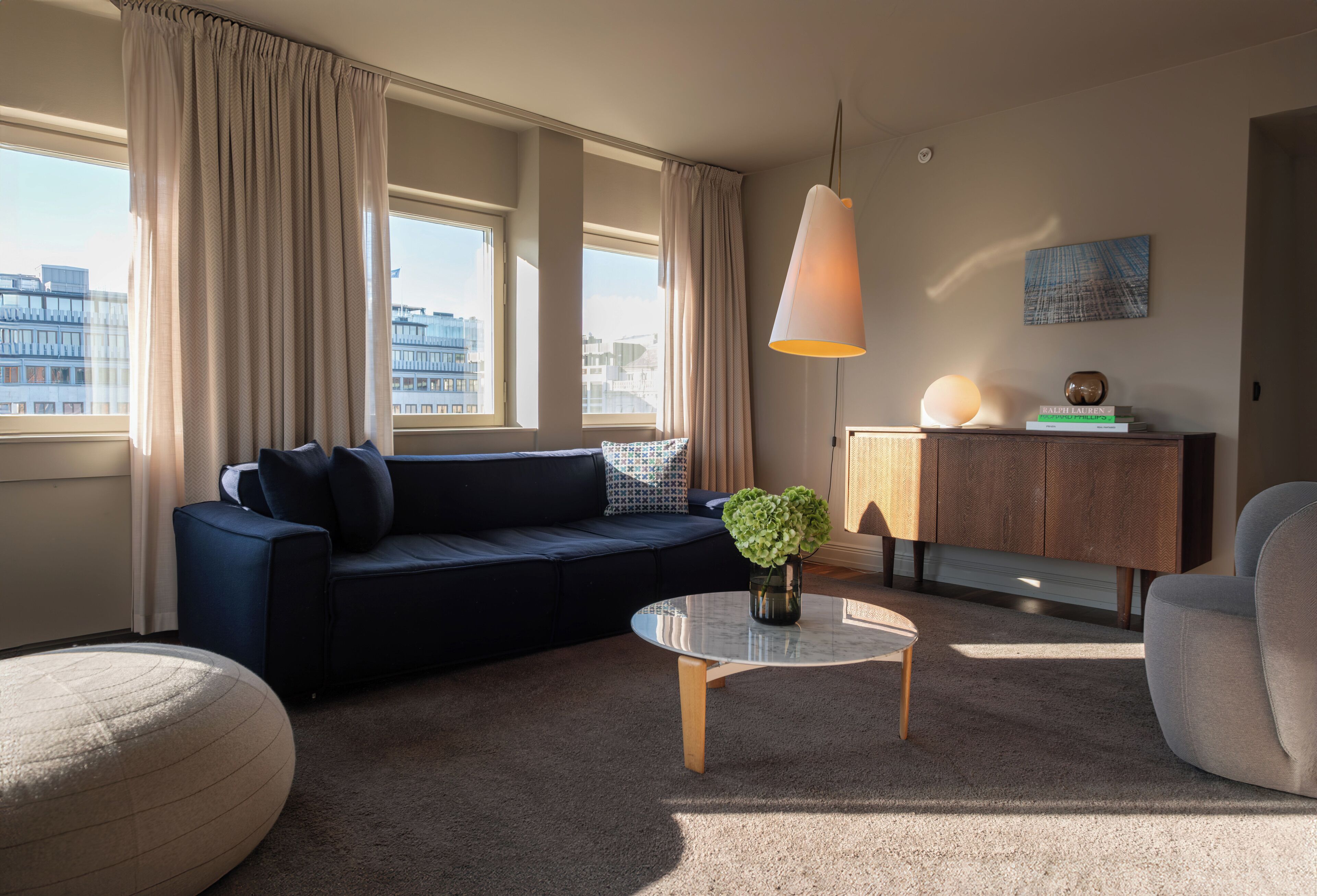 A modern living room with a dark blue sofa, a glass coffee table, and a beige ottoman. There is a wooden cabinet with decorative items and a hanging lamp.