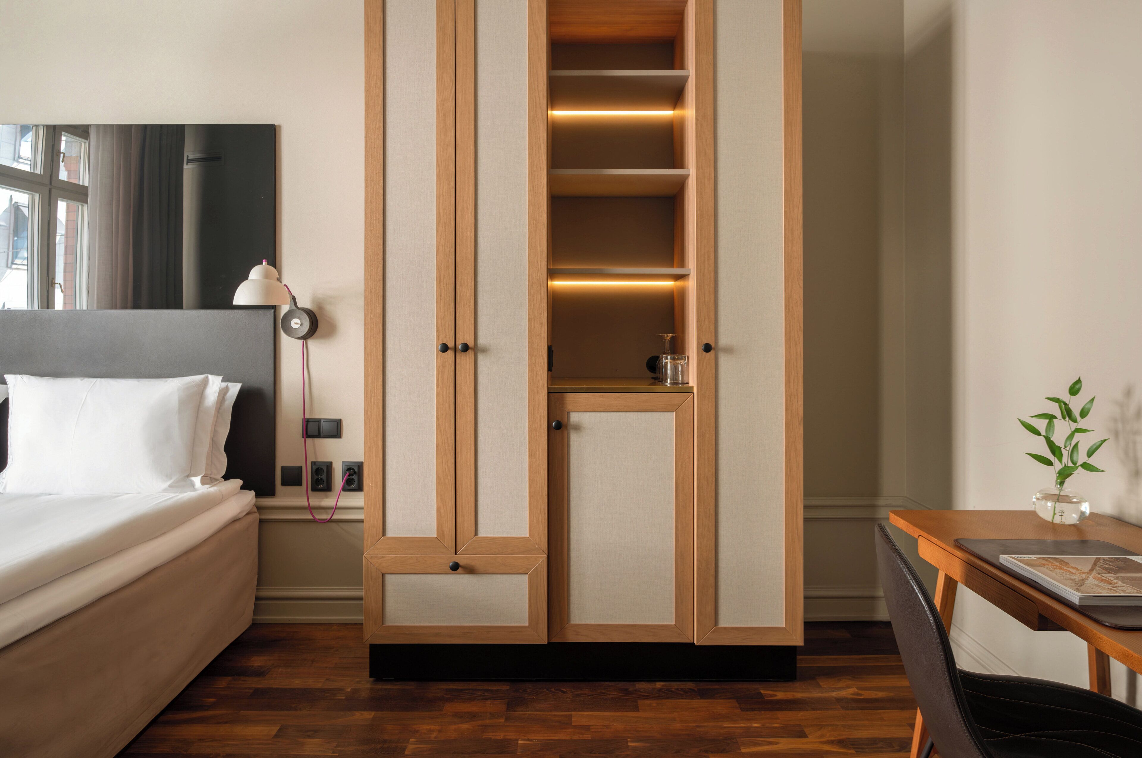 A hotel room with a bed, a desk with a chair, a wardrobe, and a bedside table with a lamp.