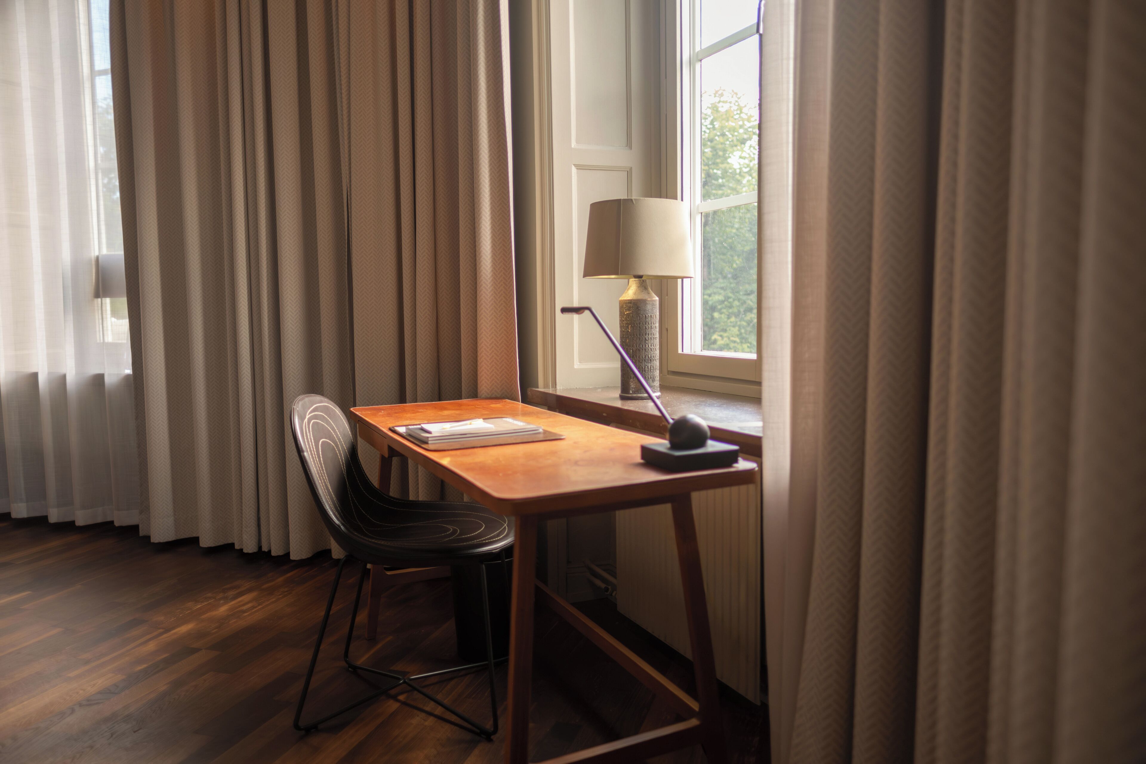 Ein Zimmer mit einem Holztisch, einem Stuhl, einer Lampe und einem Fenster mit Vorhängen.