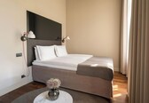 A neatly made bed with white linens, a round marble table with a vase of flowers, and a dark headboard.