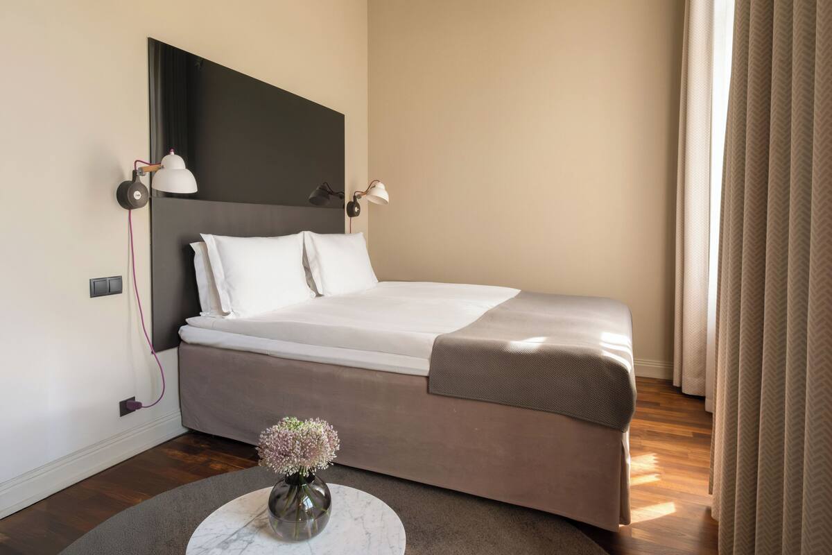 A neatly made bed with white linens, a round marble table with a vase of flowers, and a dark headboard.