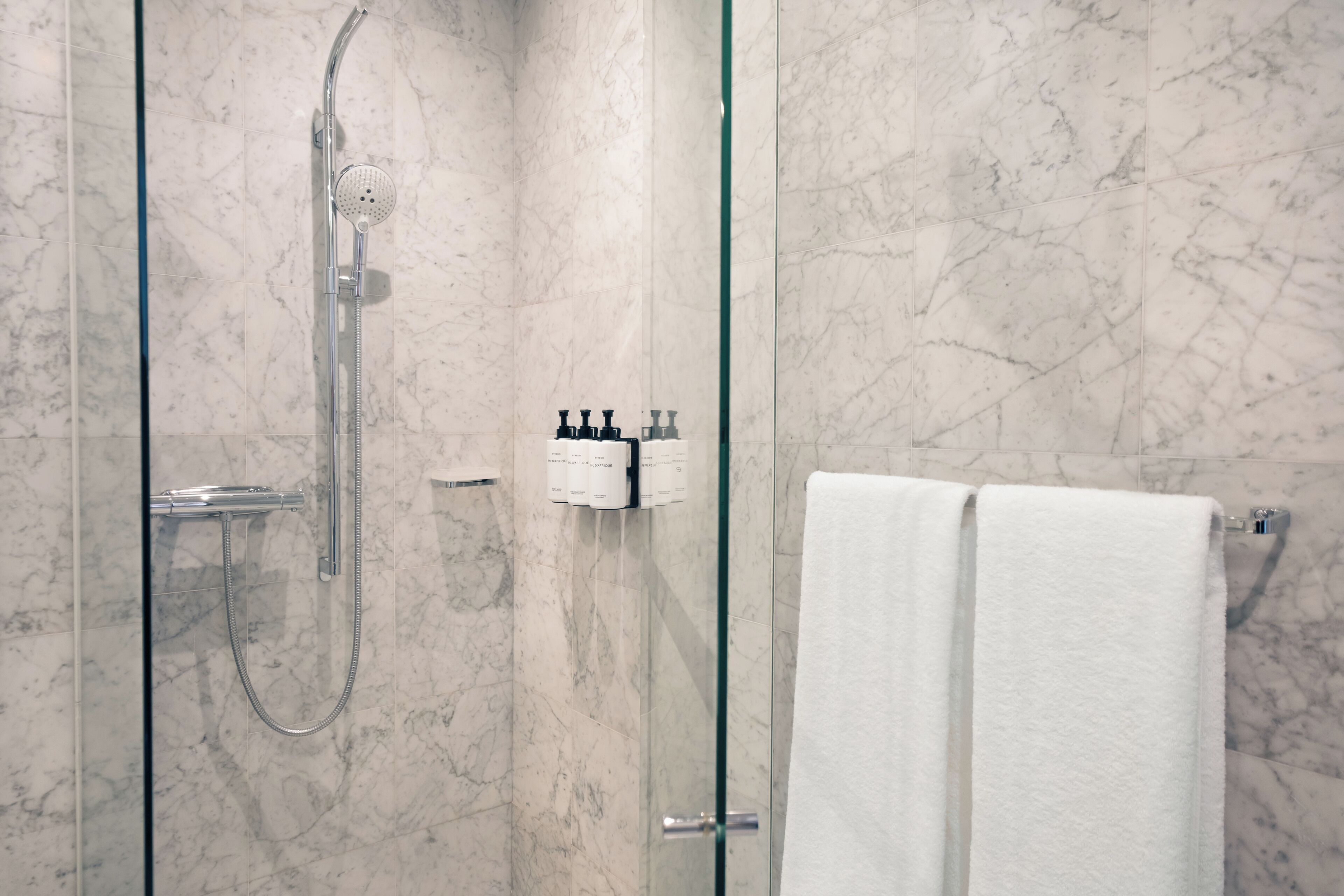 Un baño moderno con ducha envuelta en vidrio, paredes de mármol y un perchero con dos toallas blancas.