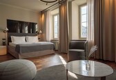 A modern hotel room with a large bed, a sitting area with a table, and a view of the city through large windows.