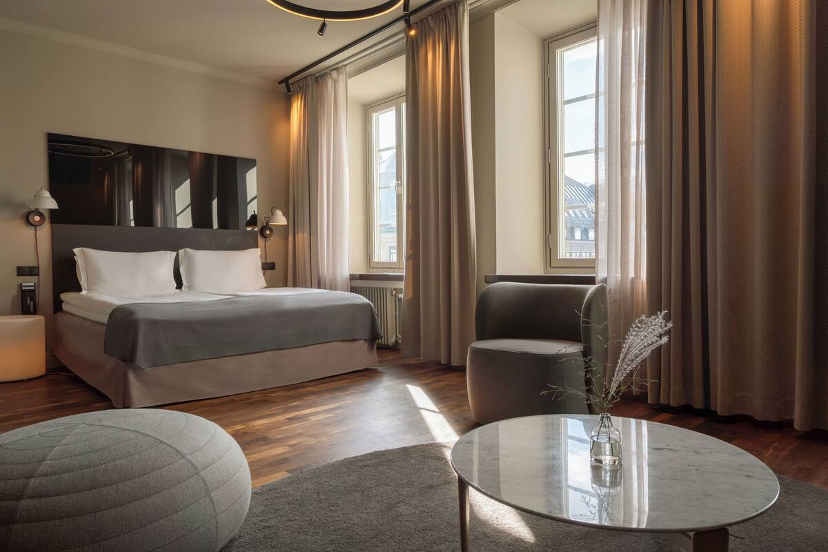 A modern hotel room with a large bed, a sitting area with a table, and a view of the city through large windows.