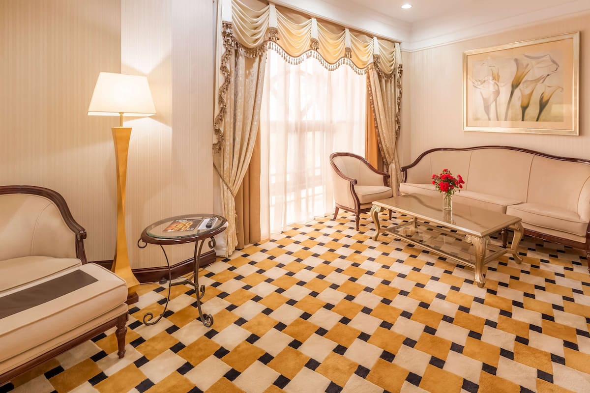 Una habitación con piso a cuadros, un sofá, una silla, una mesa con un florero y un jarrón lleno de flores, una lámpara y cortinas.