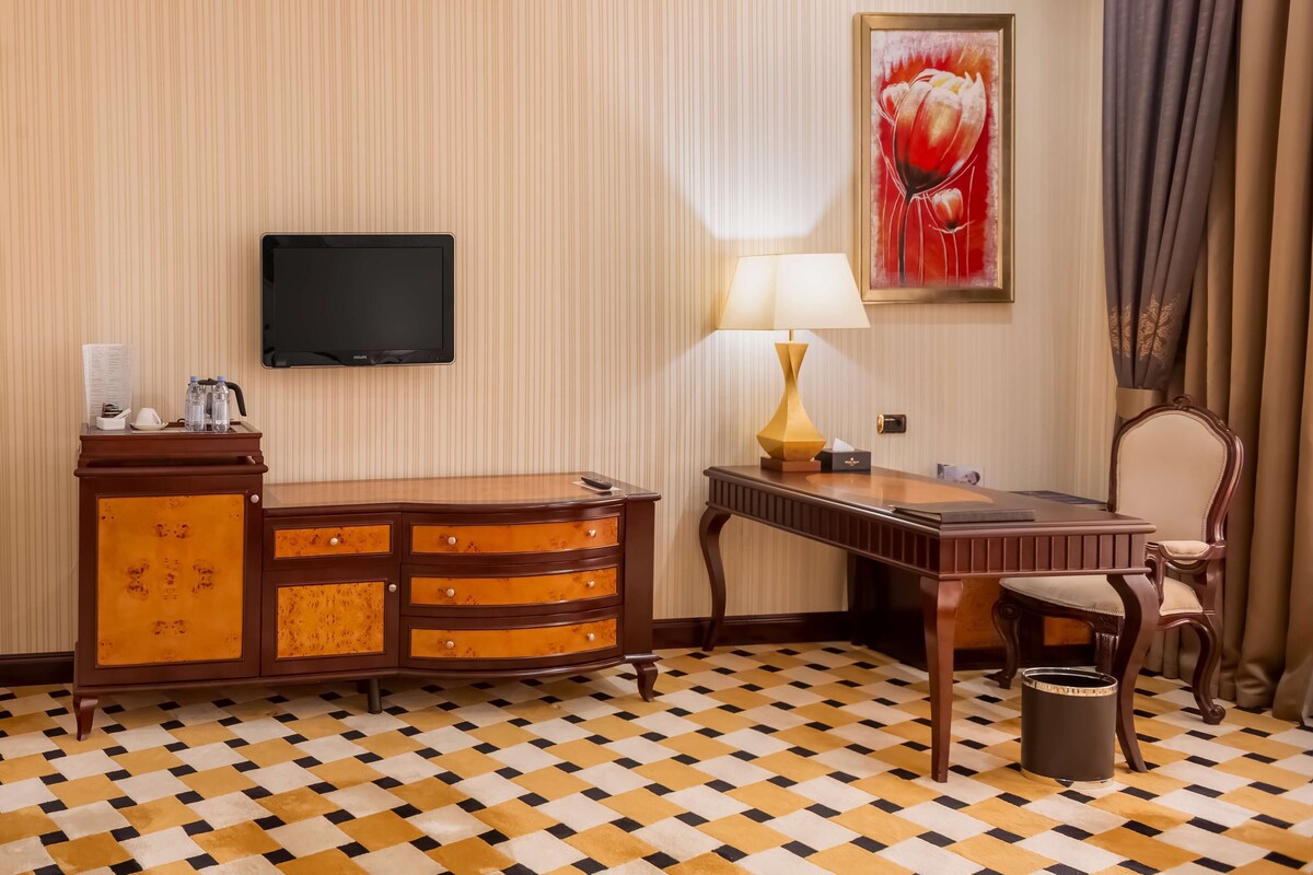 A hotel room with a checkered floor, a wooden desk with a lamp, a television, a wooden chair, and a cabinet with toiletries.
