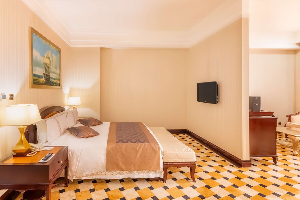 A hotel room with a large bed, a desk with a lamp, a telephone, a flat-screen TV, and a chair.