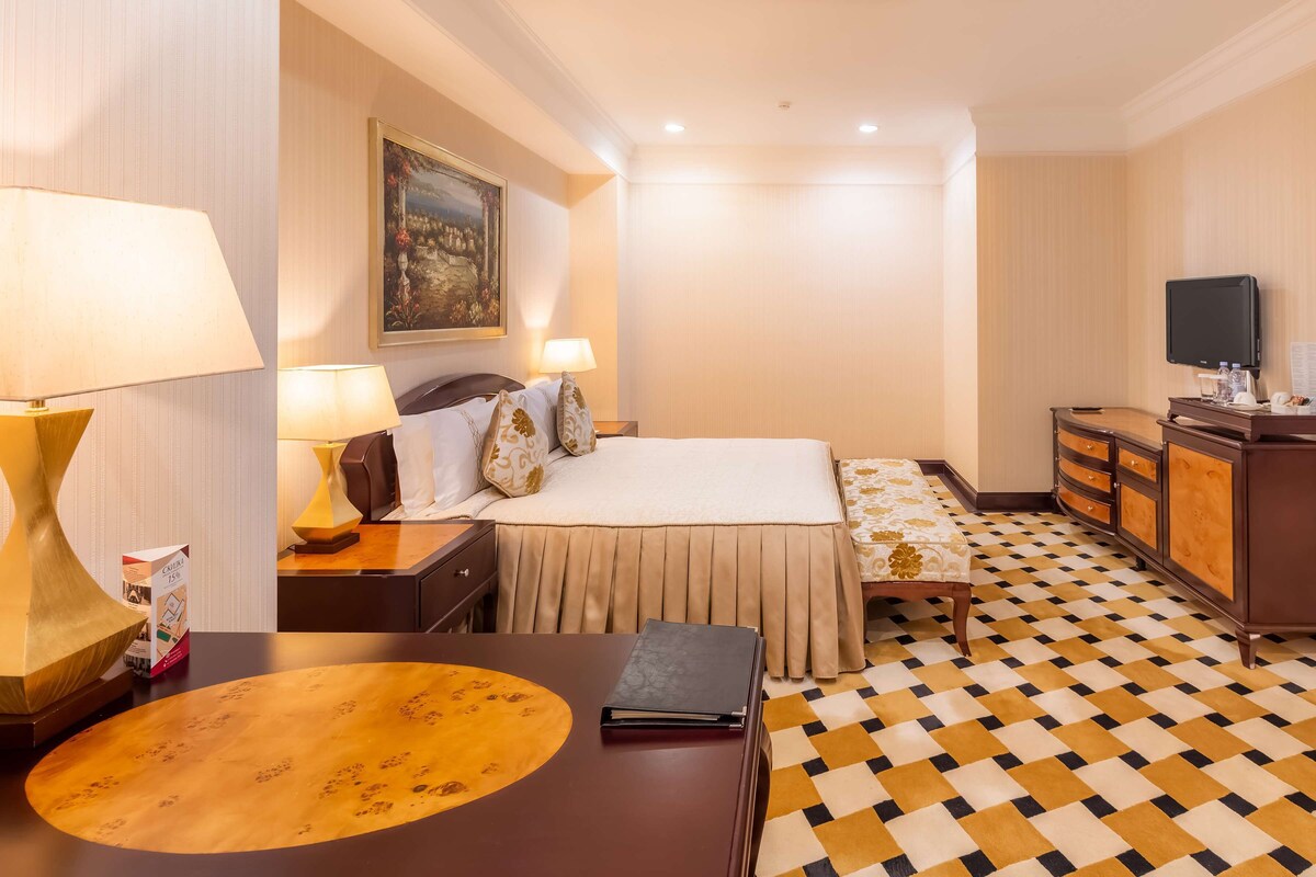 A hotel room with a bed, bedside tables, a desk, a chair, a television, and a patterned floor.