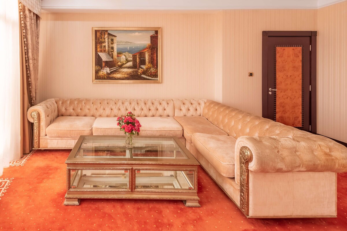 A living room with a beige sofa, a glass coffee table with a vase of flowers, and a painting of a coastal scene on the wall.