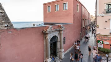 A narrow street with a pink building featuring an arched entrance, a sign for 'Rieseburg Silvester', and a view of the sea.