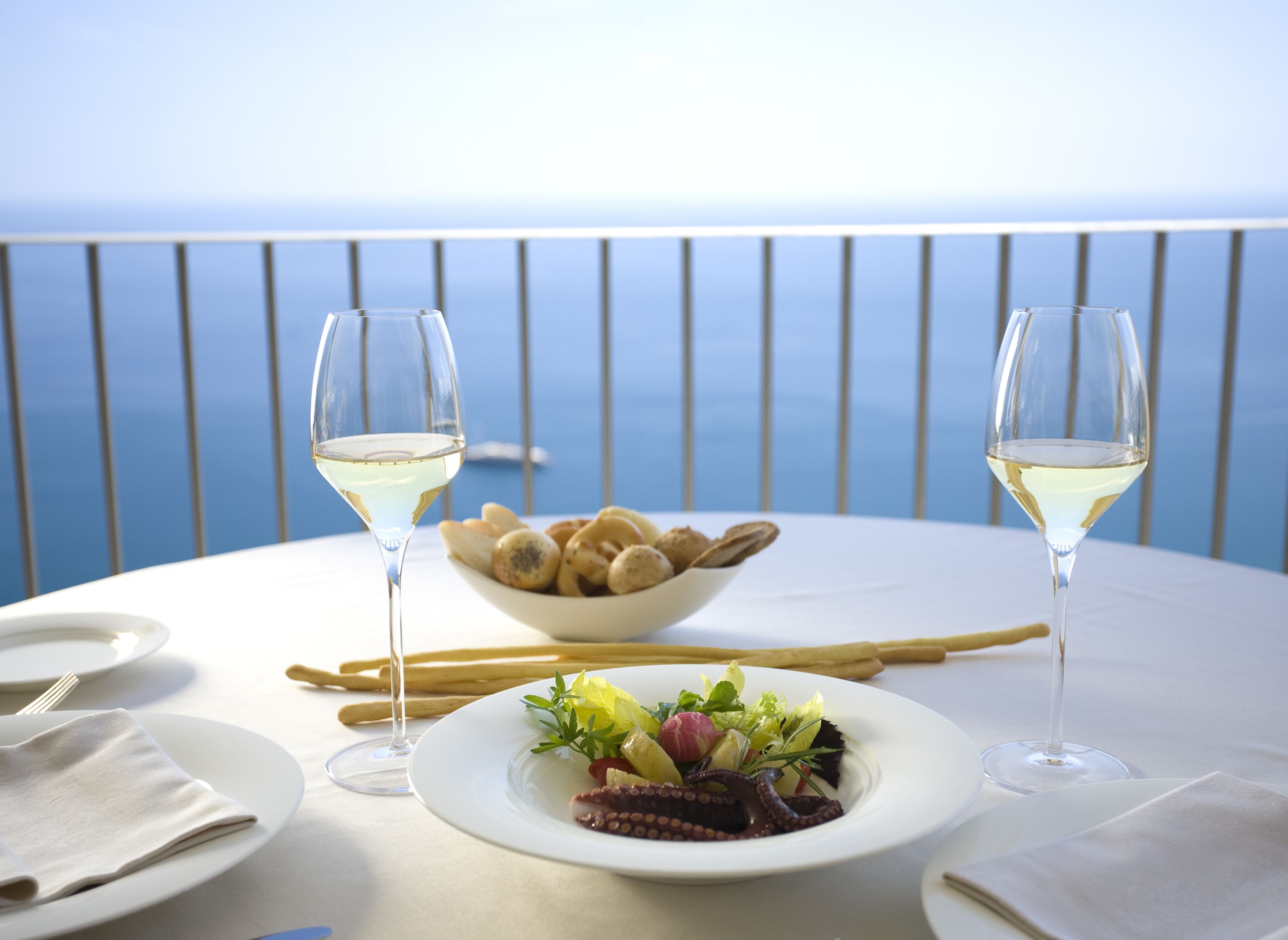 Una mesa dispuesta con una ensalada de pulpo, un cuenco de pan y dos copas de vino blanco, con vista al mar de fondo.