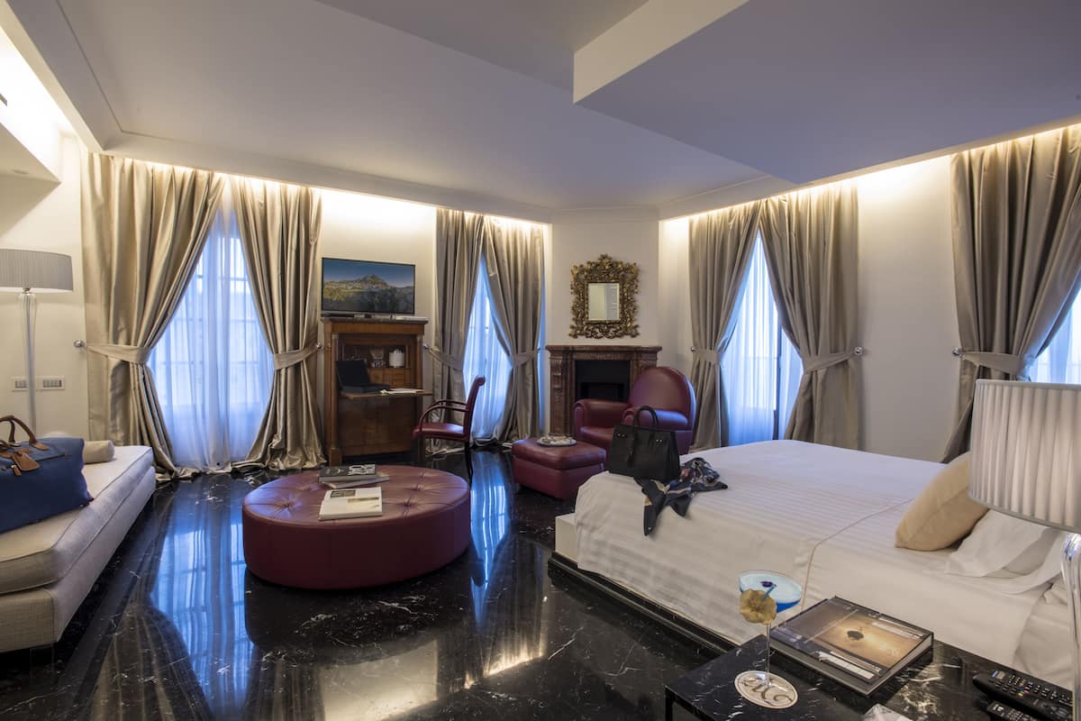 A hotel room with a bed, a seating area with a red ottoman, a fireplace, and a large mirror on the wall.