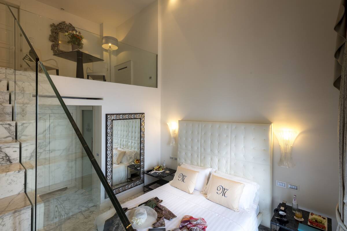 A bedroom with a marble staircase, a large bed with white linens, a nightstand with a lamp, and a mirror above the bed.
