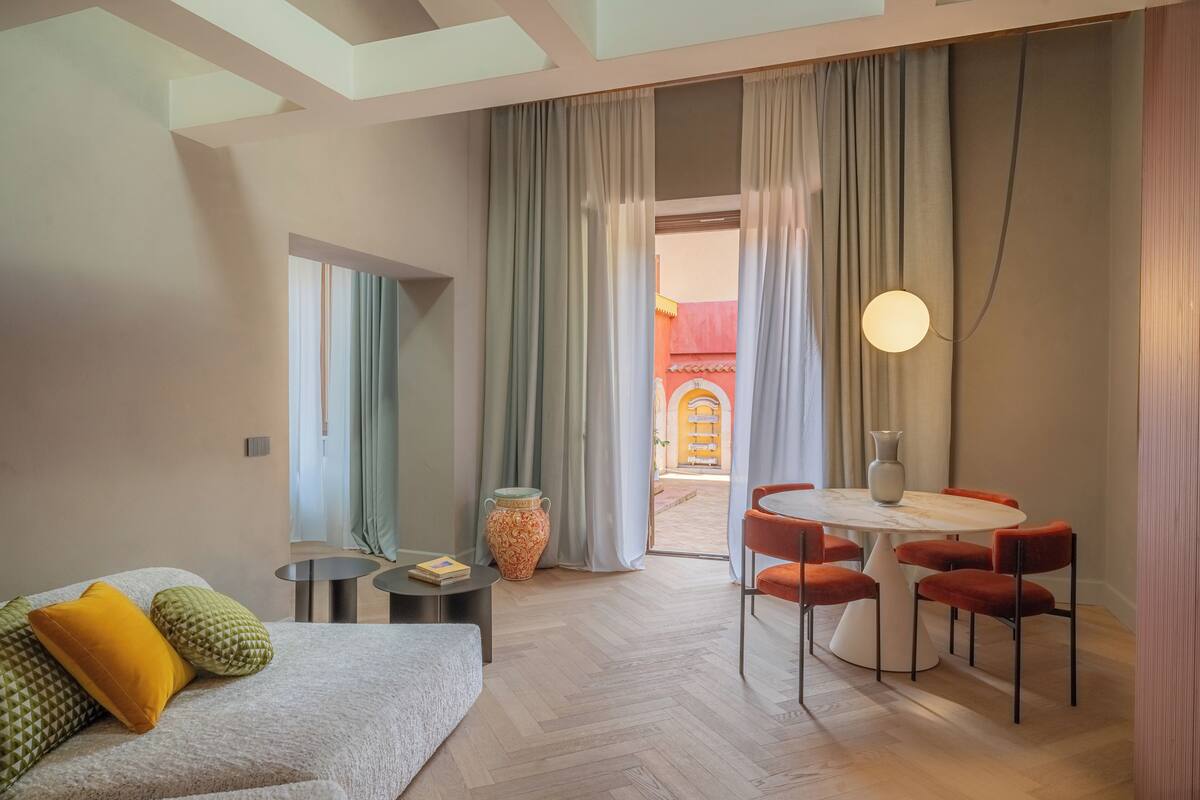 A modern hotel room with a sofa, a dining table with chairs, a vase, and a large window with curtains.