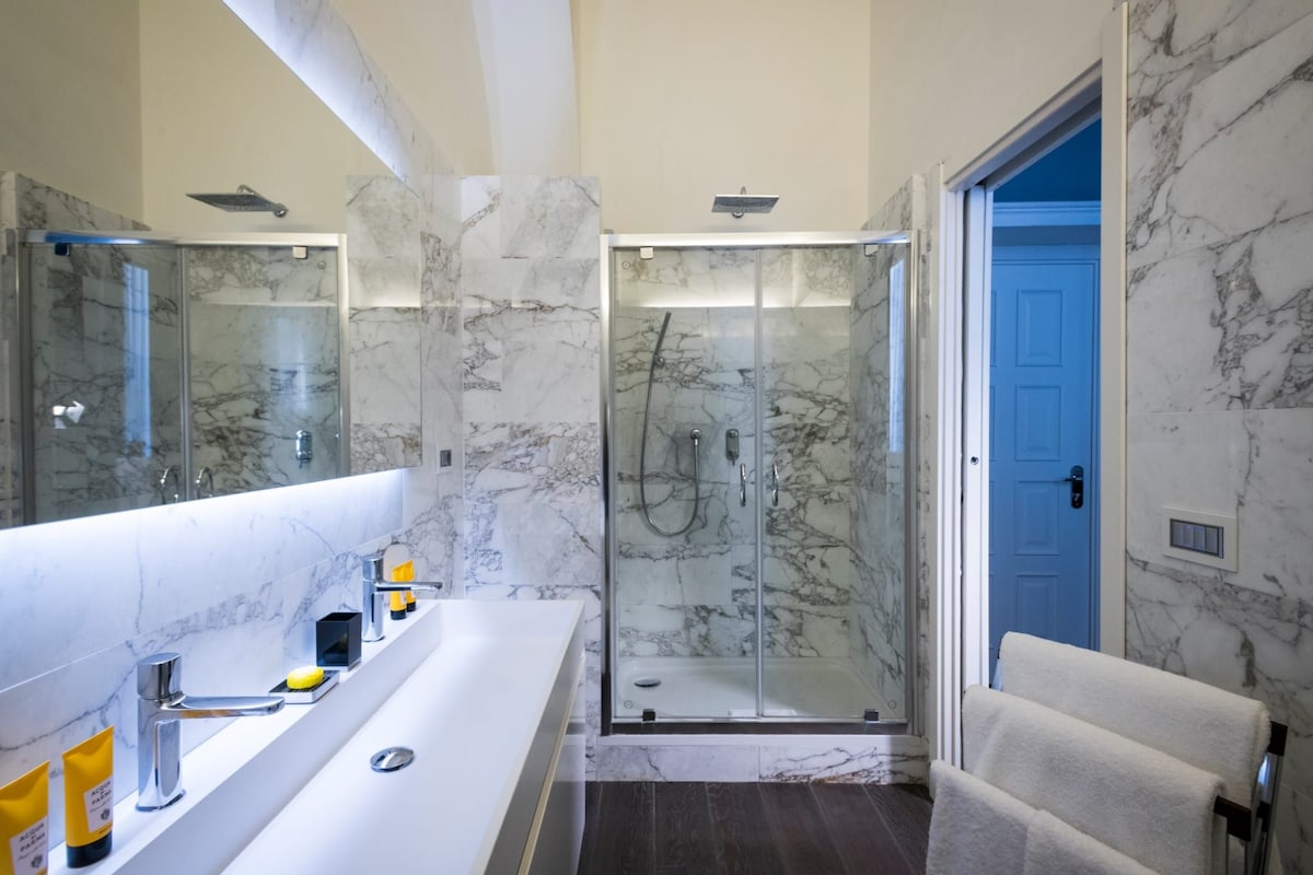 Un baño moderno con pared de mármol, cabina de ducha de vidrio y lavamanos blanco con espejo.