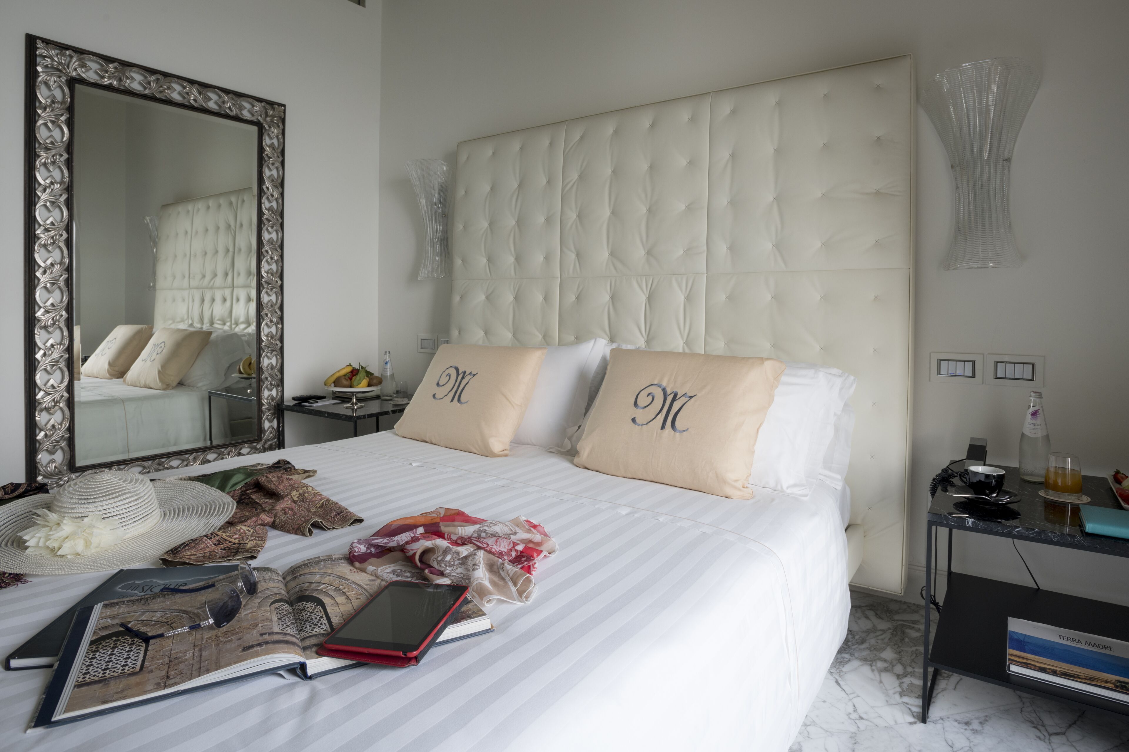 A hotel room with a large mirror, a tufted headboard, two pillows with initials, a bedside table with a phone, a book, and a tablet.