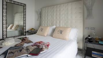 A hotel room with a large mirror, a tufted headboard, two pillows with initials, a bedside table with a phone, a book, and a tablet.