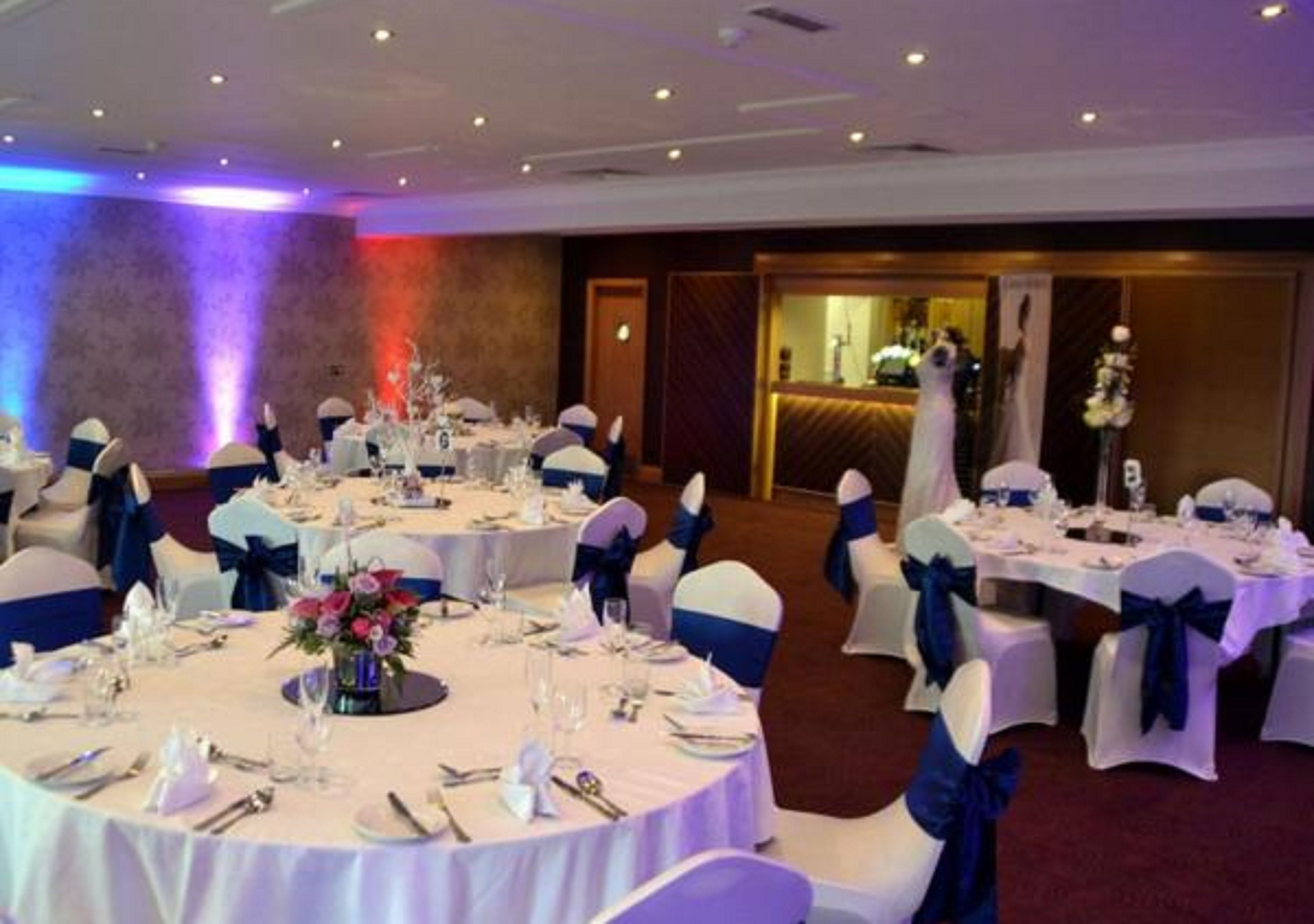 Un salón de banquetes con mesas redondas dispuestas para un evento formal, sillas blancas con cintas azules y un centro de mesa floral en cada mesa.