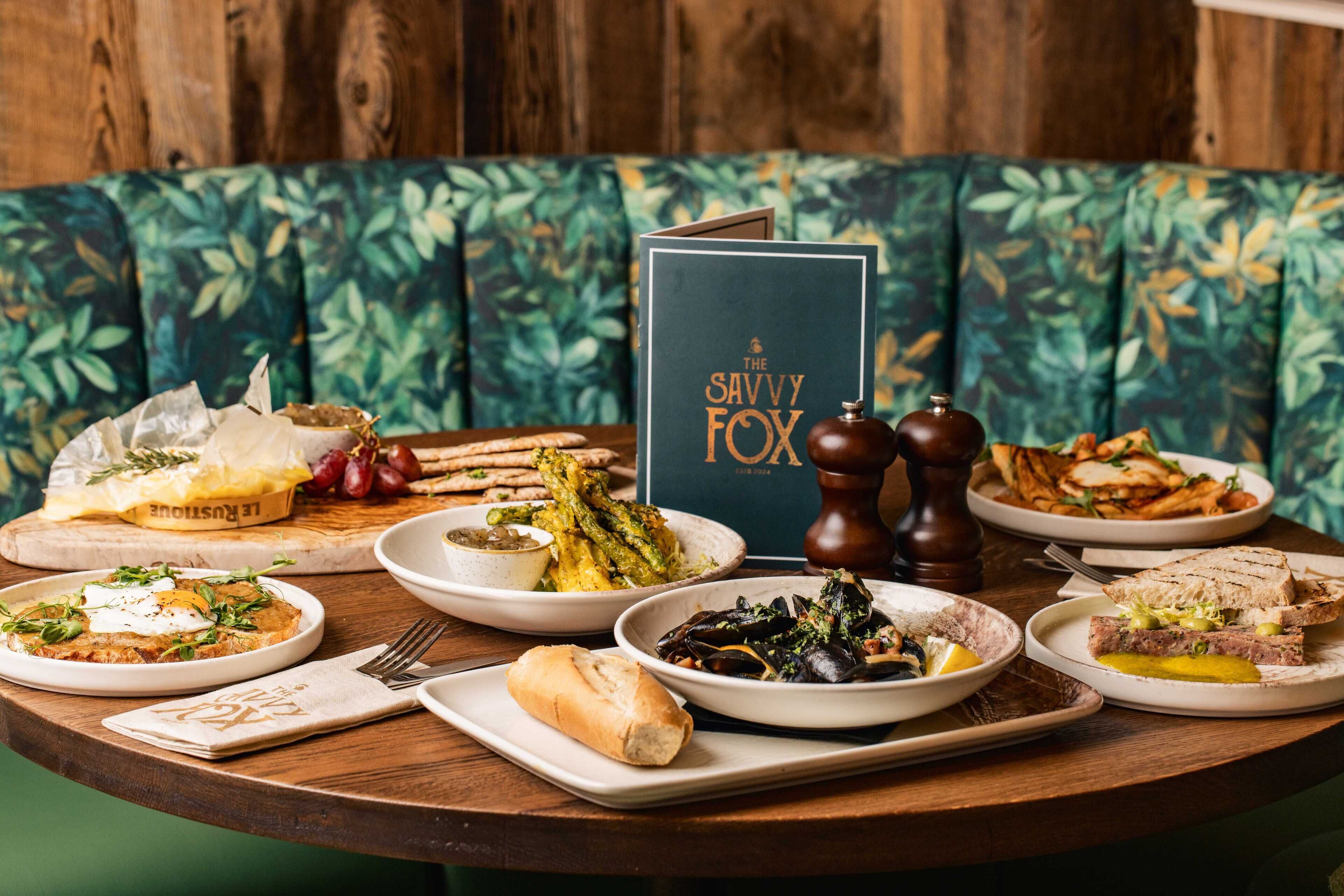 A dining table with various dishes, a menu titled "The Savvy Fox," and a wooden bench with a leafy pattern.