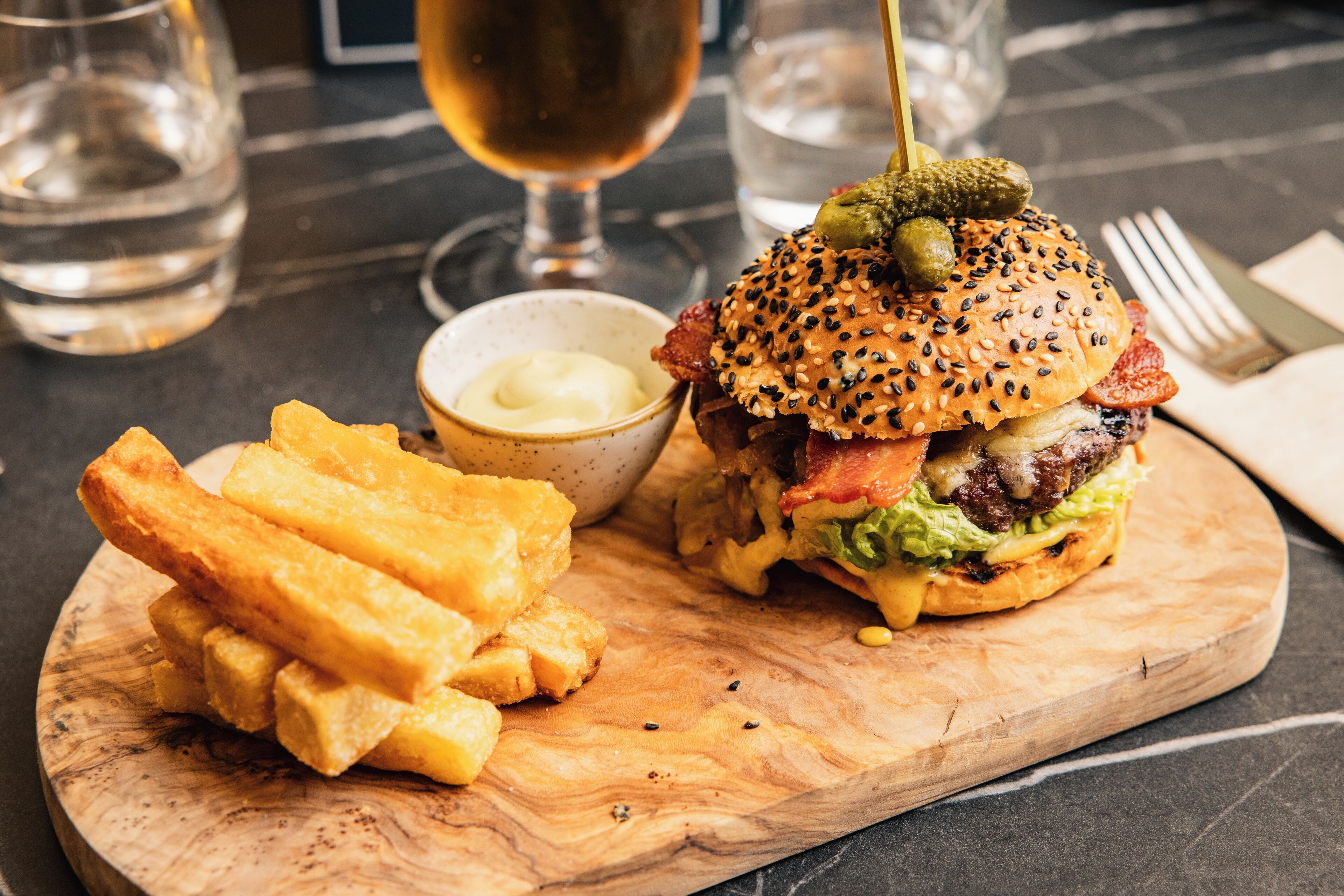 A meal consisting of a burger with various toppings and fries on a wooden board, accompanied by a small bowl of sauce.