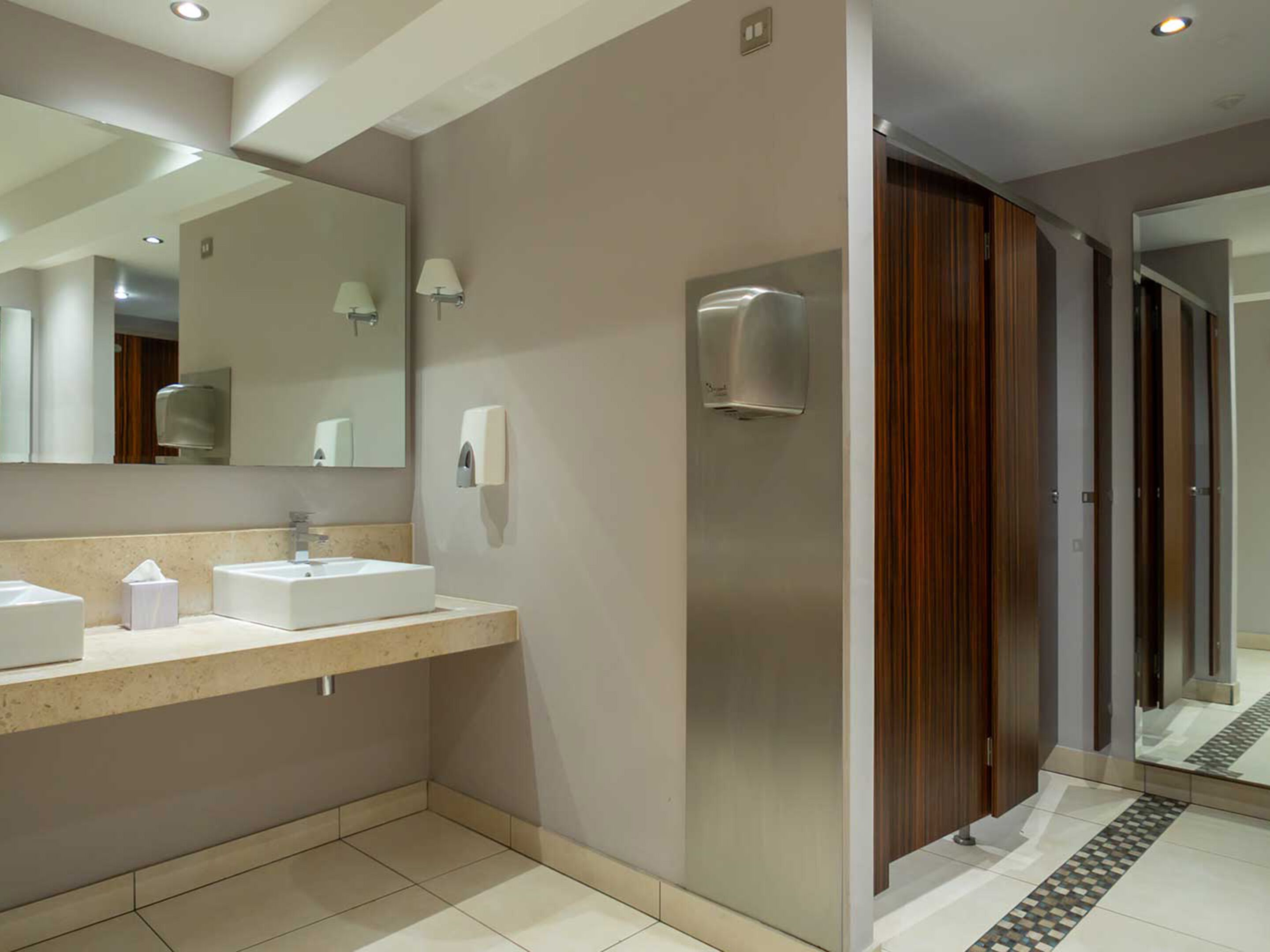A modern bathroom with a large mirror, two sinks, a towel dispenser, and a wooden door.