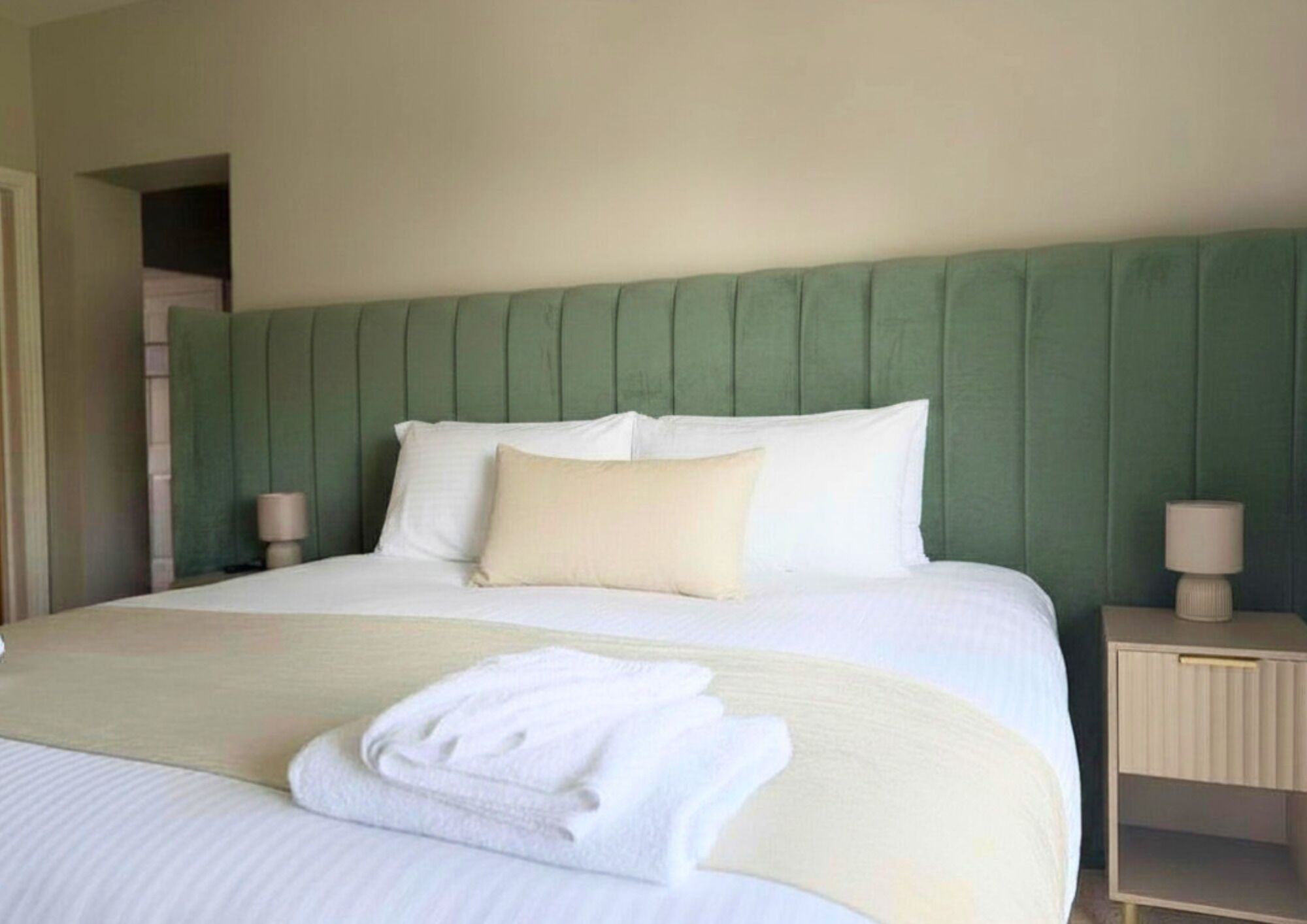 A neatly made bed with white linens, a green upholstered headboard, and a bedside table with a lamp.