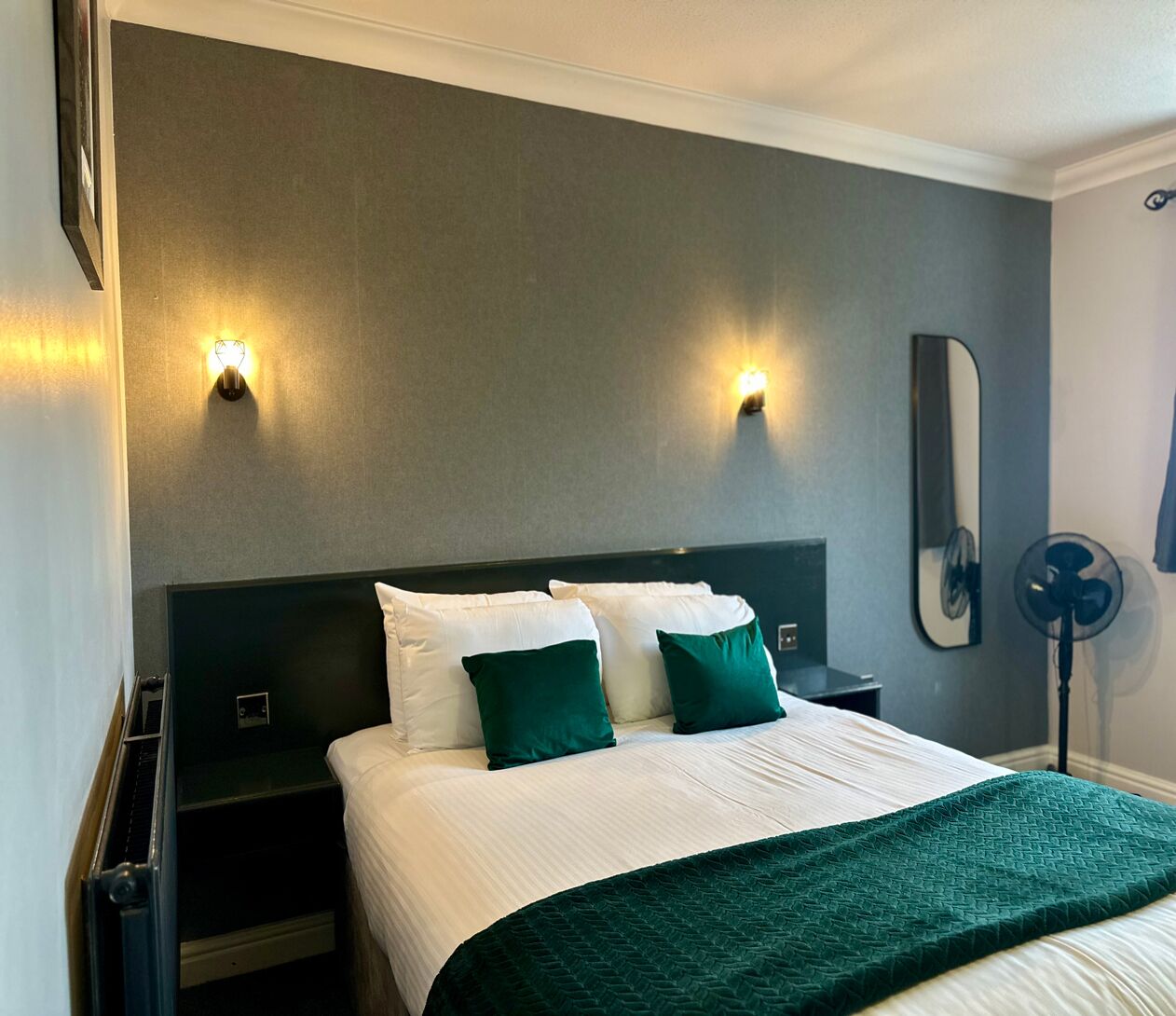 Una habitación de hotel con una cama, dos mesitas de noche, un espejo en la pared y un ventilador de pie.