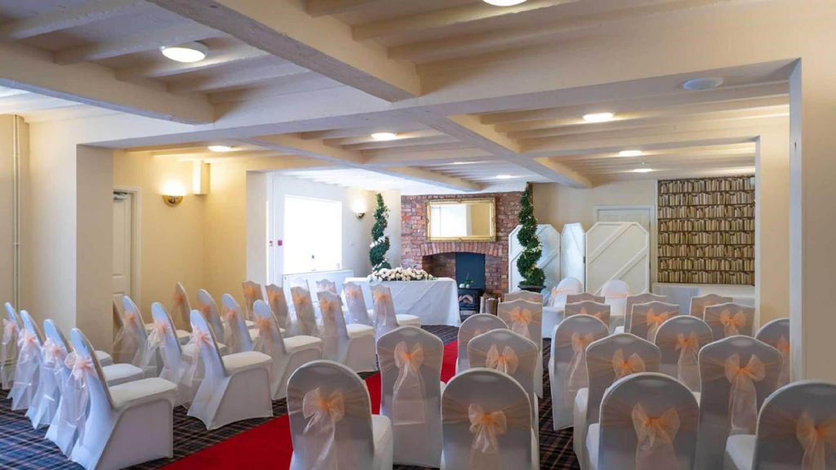 Un salón de conferencias con filas de sillas blancas adornadas con lazos naranjas, una chimenea y una estantería.