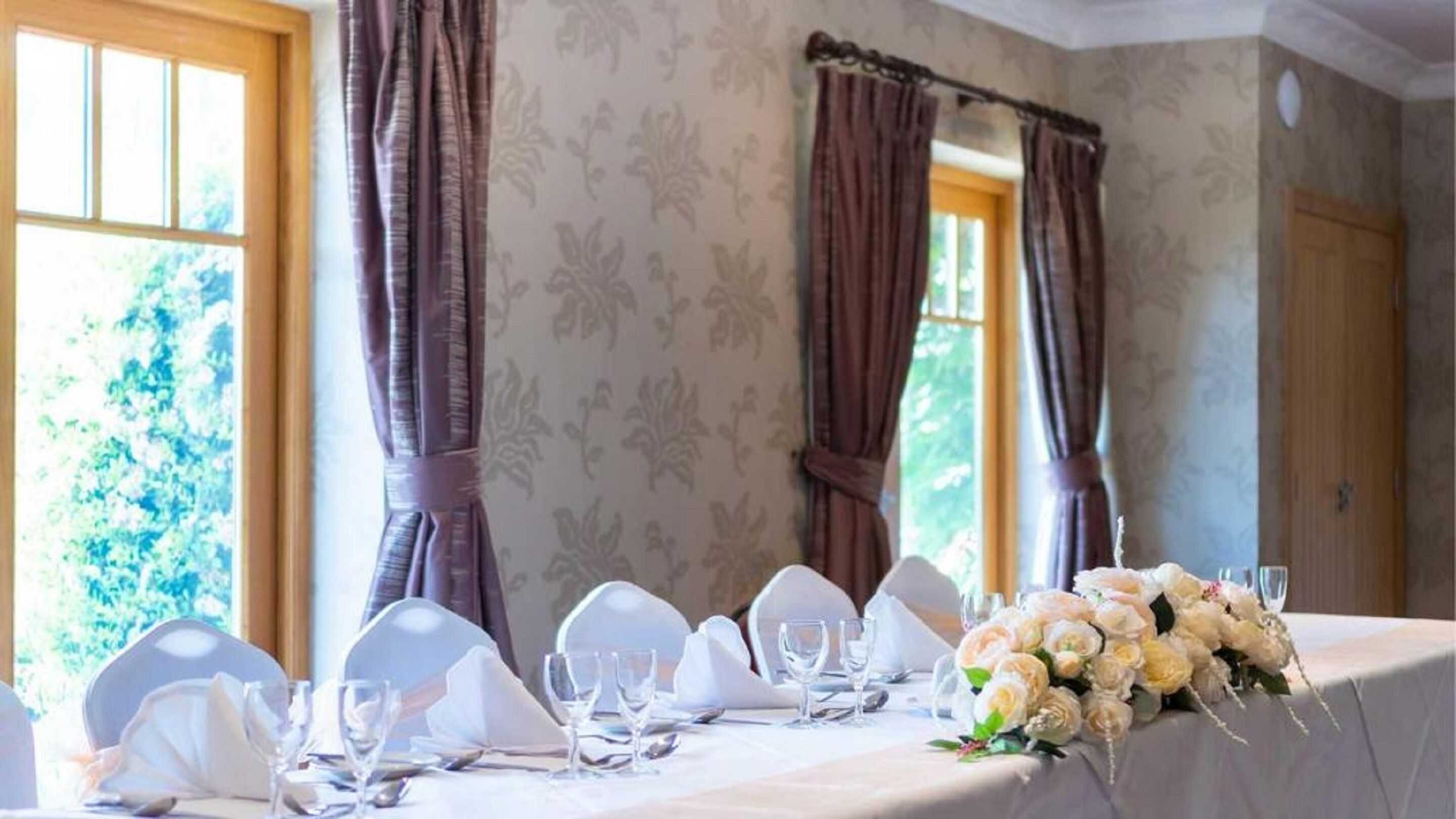 A table set for a formal event with white chairs and table settings, floral centerpieces, and large windows with curtains.