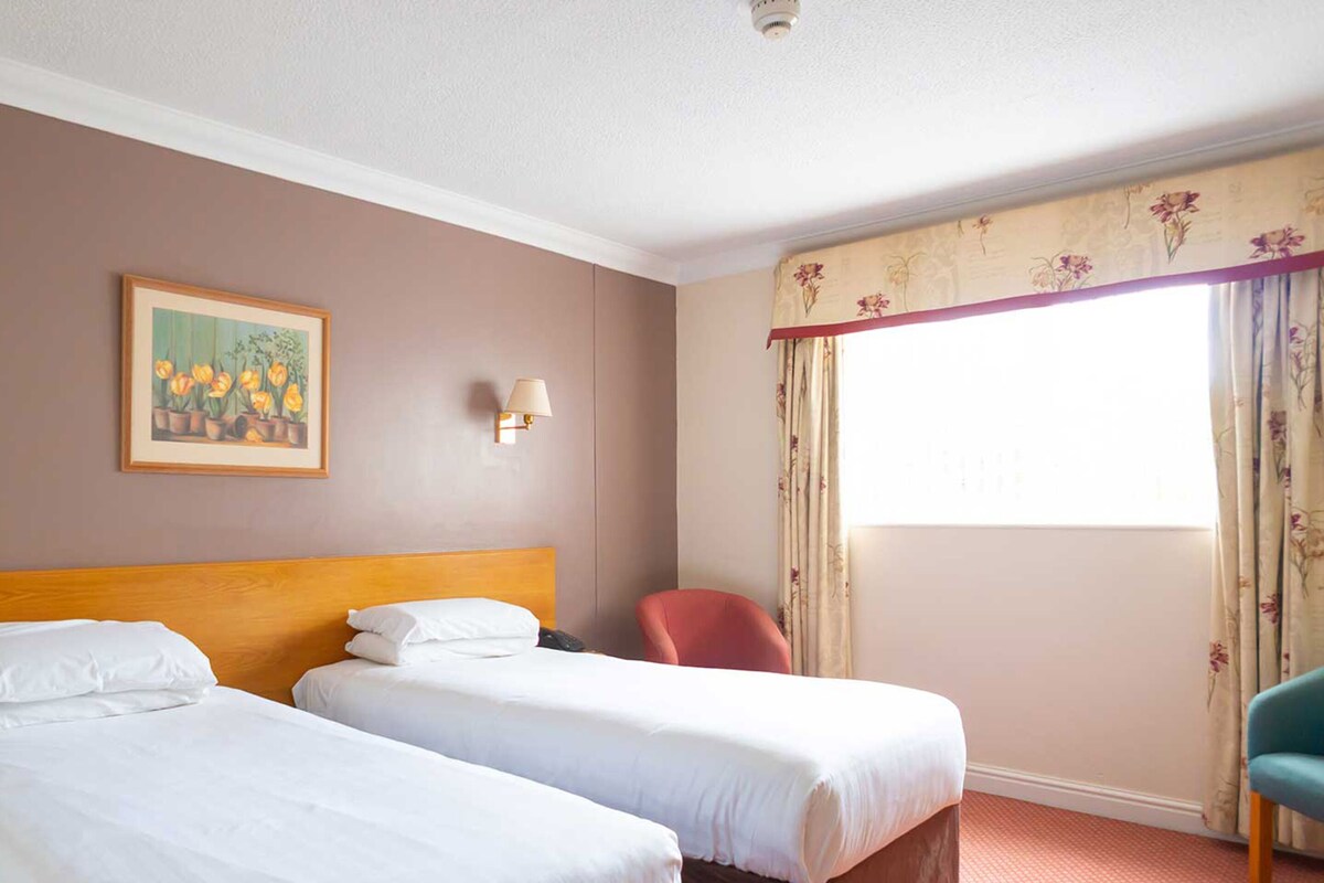 Quarto de hotel com duas camas, uma cadeira, um quadro, uma luminária e uma janela com cortinas.