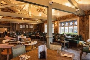 A restaurant with wooden interiors, multiple dining tables and chairs, and a large window with diamond-shaped panes.