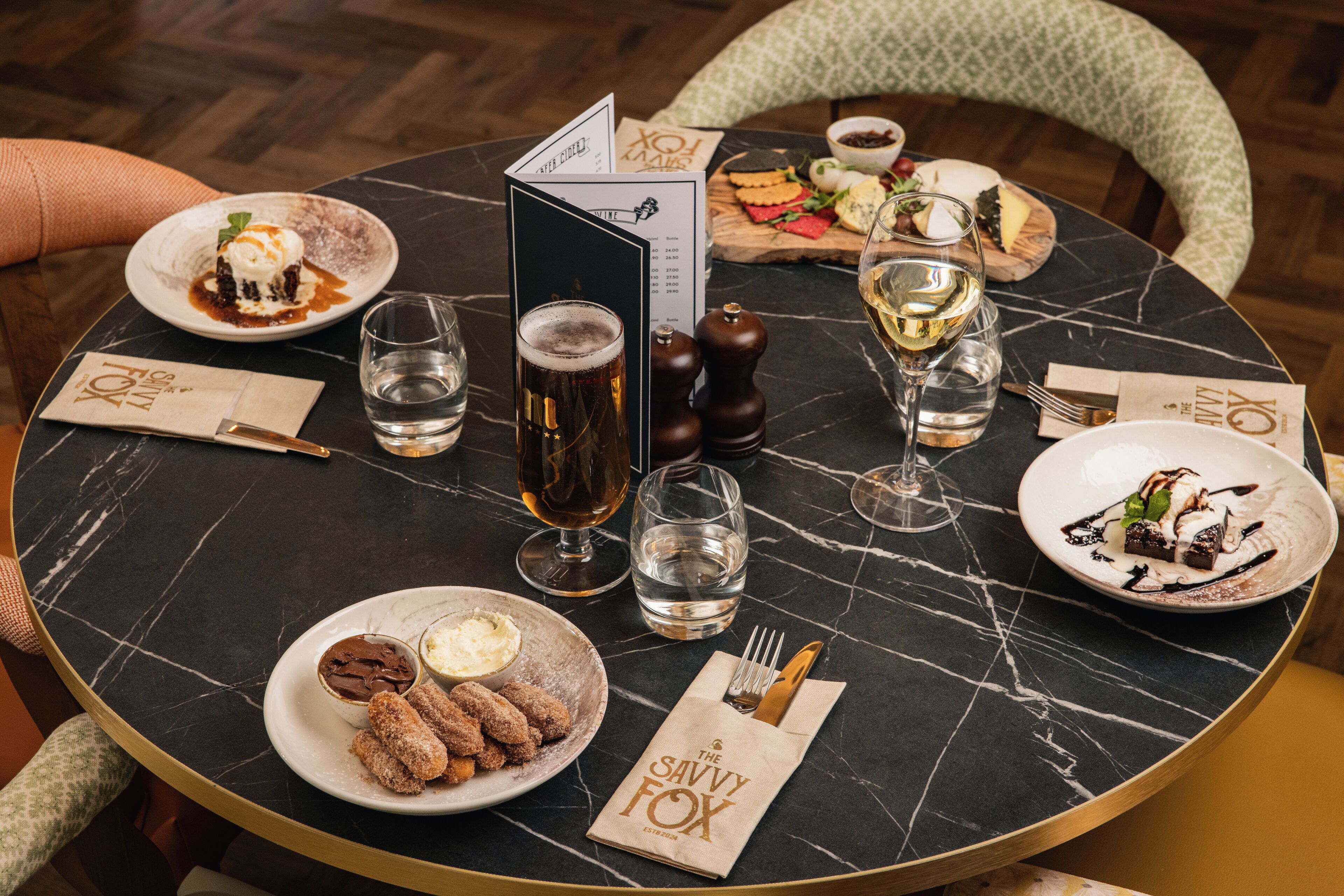 A round table with a black marble top, surrounded by chairs. There are plates of food, glasses of wine, and a menu.