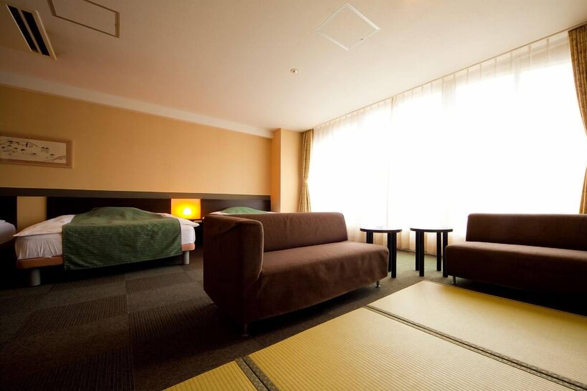 A hotel room with a bed, two sofas, a small table, and a large window with curtains.