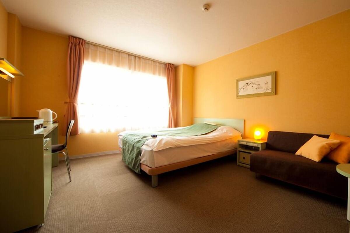 A hotel room with a bed, a sofa, a desk with a kettle, a nightstand with a lamp, and a window with curtains.