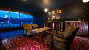 A cozy lounge area with a large window offering a view of the cityscape and mountains. There are plush chairs, a wooden table, and a decorative cabinet.