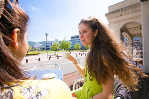 A person holding a cup at a table with a view of a street and buildings in the background.