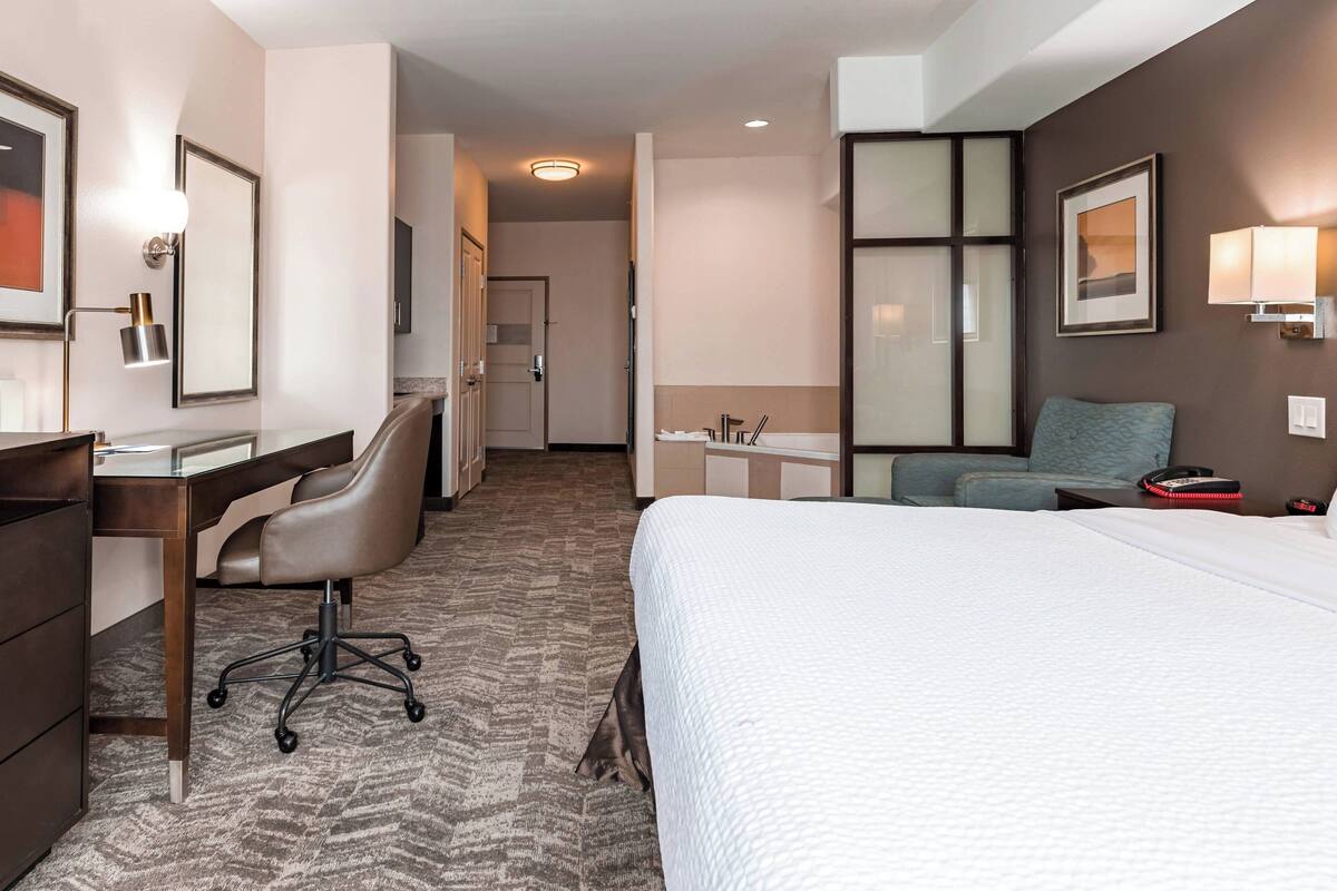A hotel room with a bed, desk, chair, and a small table with a lamp.