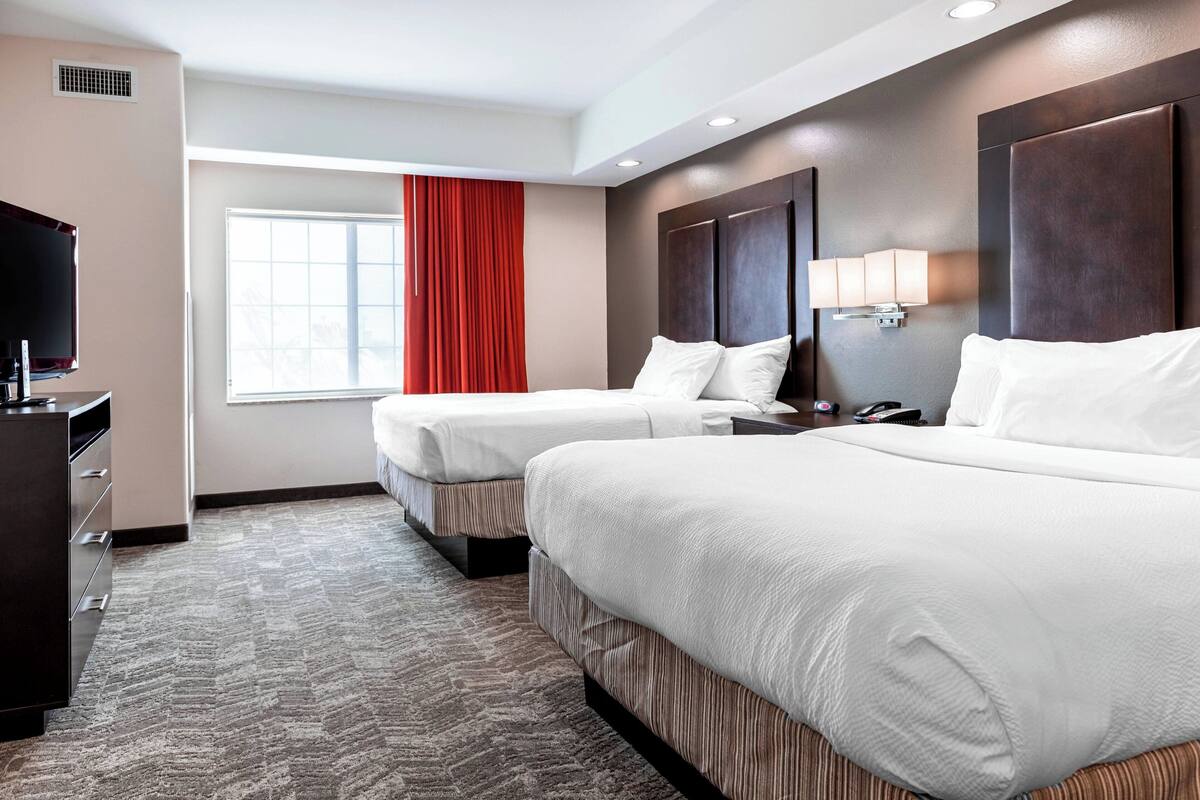 A hotel room with two beds, a TV, a desk, and a window with red curtains.