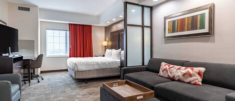 A hotel room with a bed, a gray sofa, a wooden side table, a desk with a chair, a TV, and a window with red curtains.