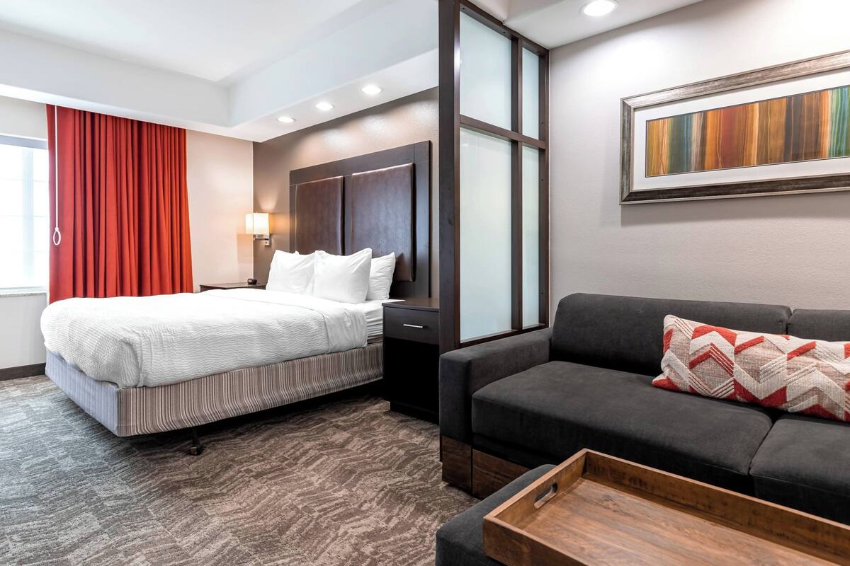 A hotel room with a bed, a sofa, a wooden side table, a wall-mounted lamp, and a large window with curtains.