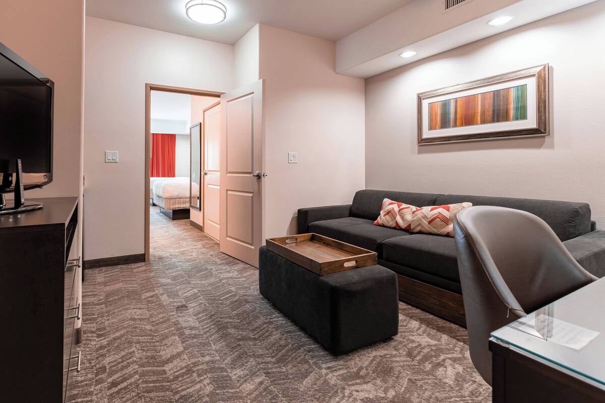 A modern hotel room with a sofa, ottoman, and a flat-screen TV.
