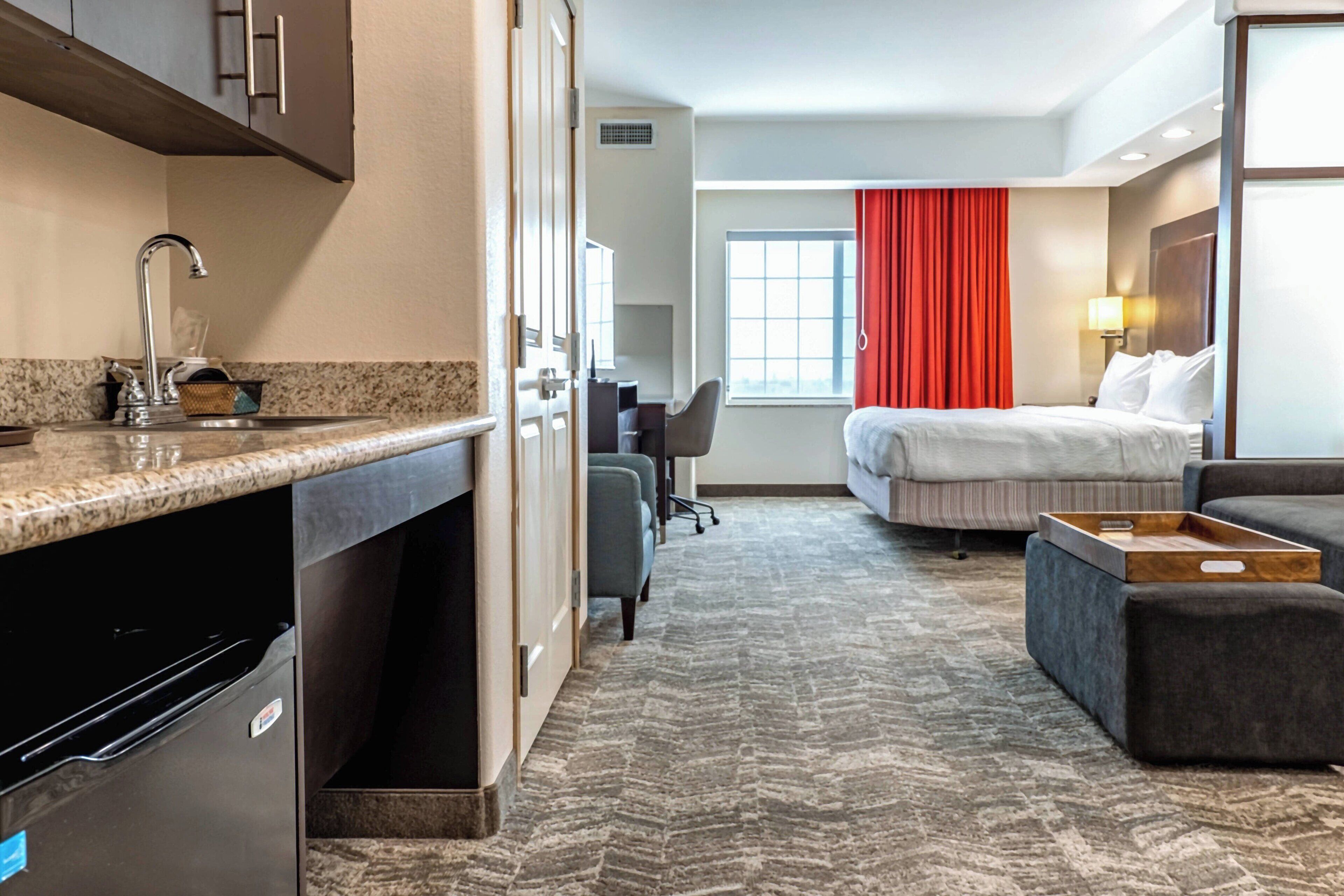 A hotel room with a kitchenette, a bed, a desk, and a window with red curtains.