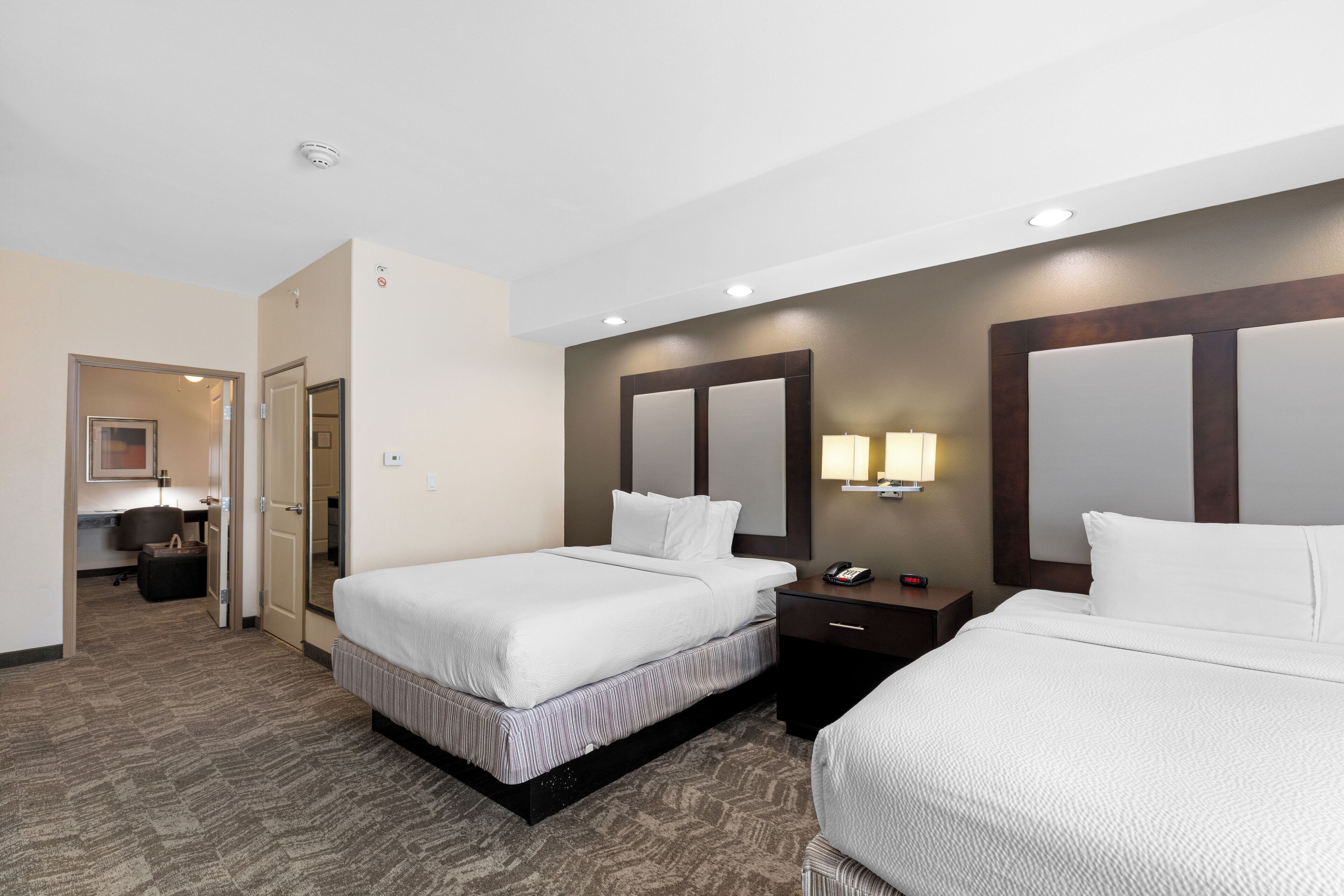 Una habitación de hotel con dos camas, un escritorio, una silla, una lámpara y un espejo.